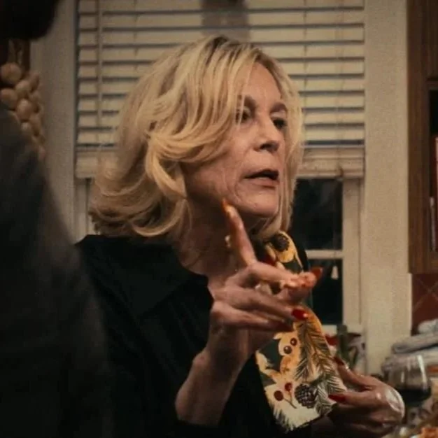 An elderly woman with blonde hair, wearing a patterned blouse, is gesturing with her hand while speaking indoors near a window with blinds. Jamie Lee Curtis, The Bear.