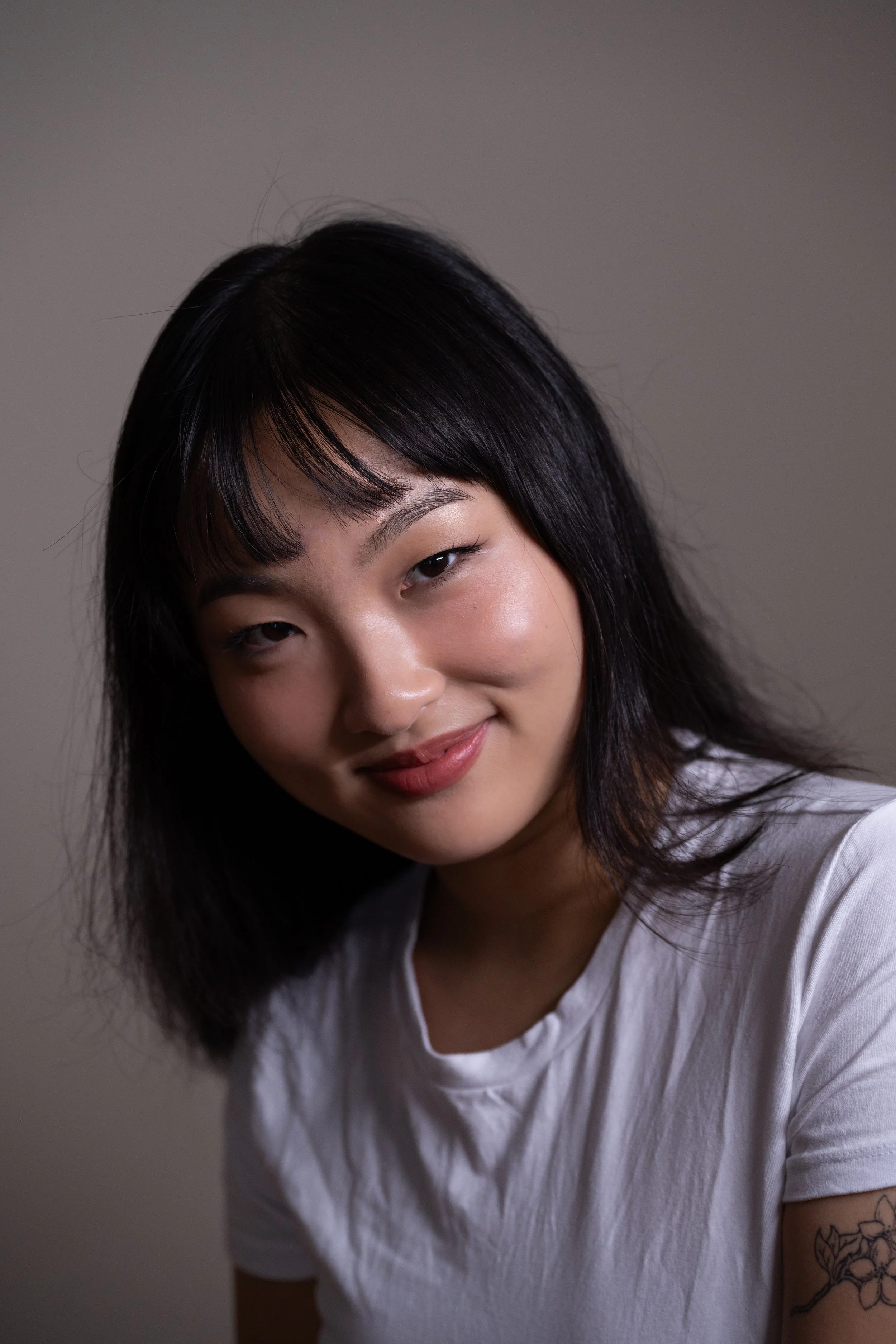 Kasey Lim, graphic design artist. Art Director of "The Tooth Fairy" short film. A woman with shoulder-length black hair, wearing a white t-shirt, smiling slightly, with a tattoo of flowers on her upper arm, against a plain gray background.