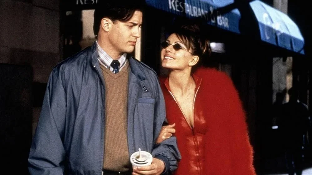 A young man and woman standing close together outdoors. The woman, wearing sunglasses and a red jacket, appears to be talking happily to the man, who is looking down with a neutral expression, holding a cup. Bedazzled, Elizabeth Hurley 