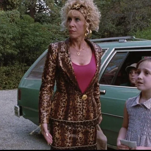 A woman with curly blonde hair wearing a leopard print suit stands outdoors near a green station wagon. A young girl with a gray dress and short hair is in the background, holding a piece of paper. Rhea Pearlman, Matilda.