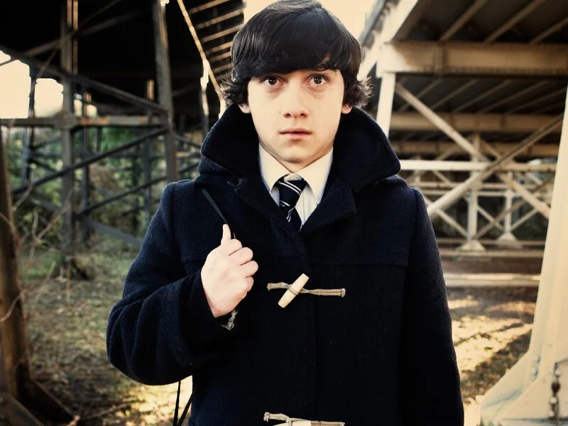 Young man with dark hair, wearing a black coat, white shirt, and striped tie, standing outdoors under a bridge or structure, carrying a bag over his shoulder, with trees in the background. Submarine. 