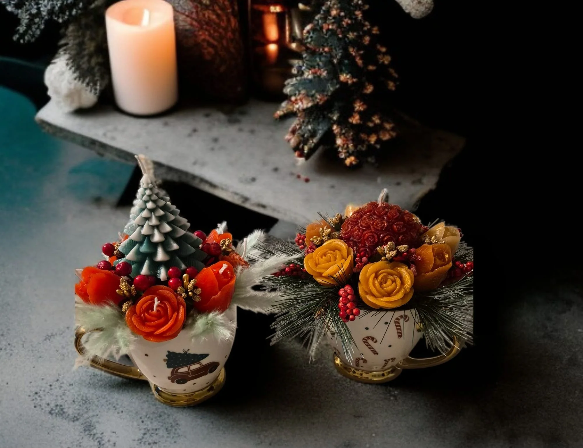 Two Christmas-themed floral arrangements in cups, featuring roses, berries, pine branches, and holiday decor, on a gray surface with a decorated Christmas tree and candles in the background.