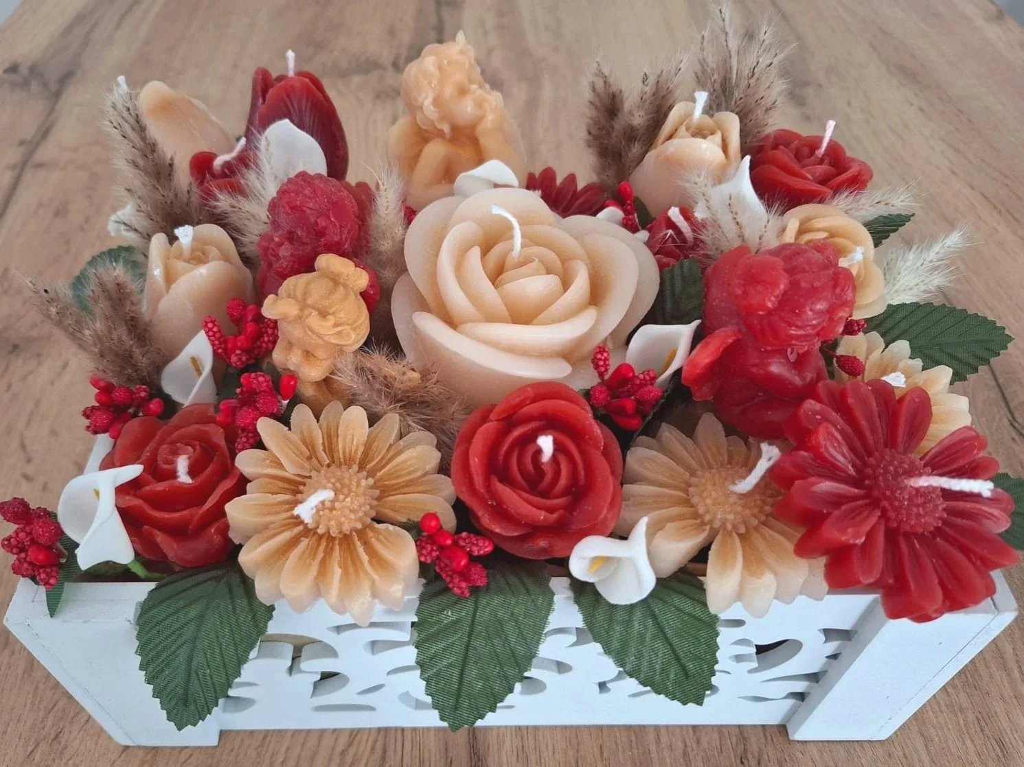 A decorative arrangement of candles shaped like flowers in a white wooden basket, with candles resembling roses, daisies, and other flowers in red, beige, and cream colors, accented by green leaves and small red berries.