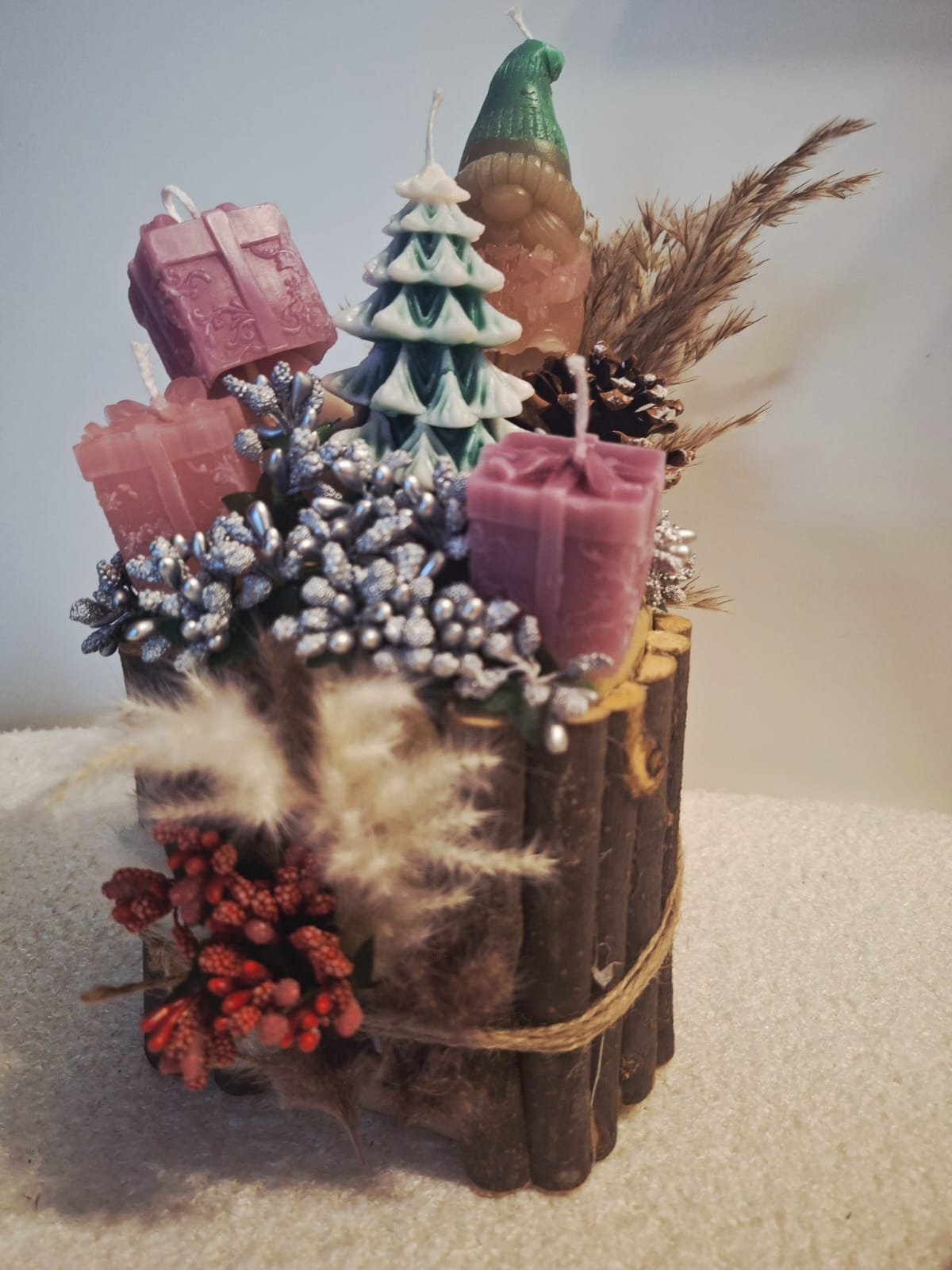 Decorative wooden log with candles, artificial pinecones, dried plants, and holiday-themed ornaments including a green candle, pink candles, and a small Christmas tree figure.