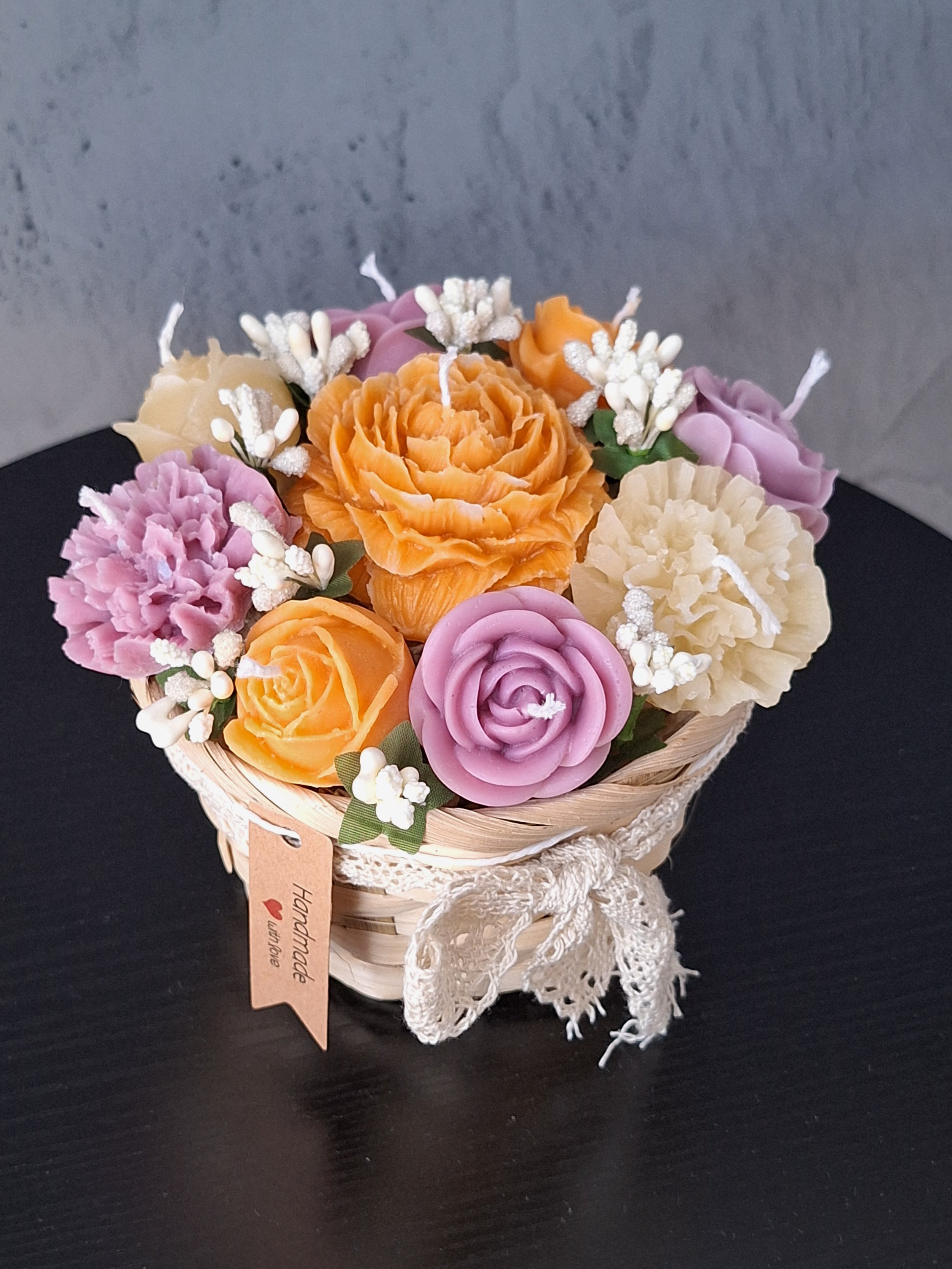 A small decorative basket filled with colorful soap flowers in shades of orange, pink, purple, and cream, decorated with white artificial small flowers and greenery, tied with a lace ribbon.