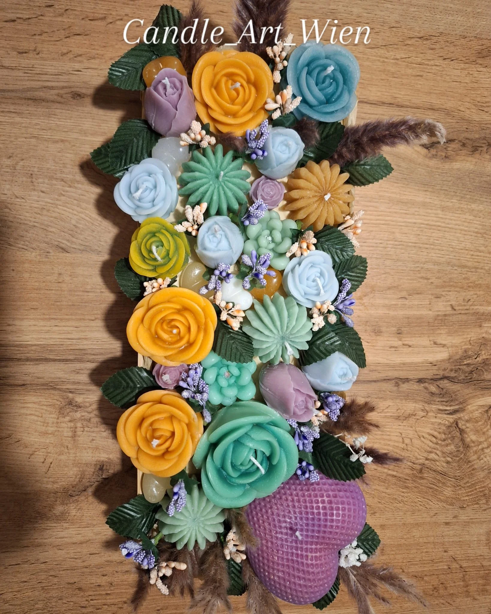 Bunte Kerzen in Blumenform in verschiedenen Farben, umgeben von grünen Blättern, kleinen Blumen und Federn, auf einem Holztisch.