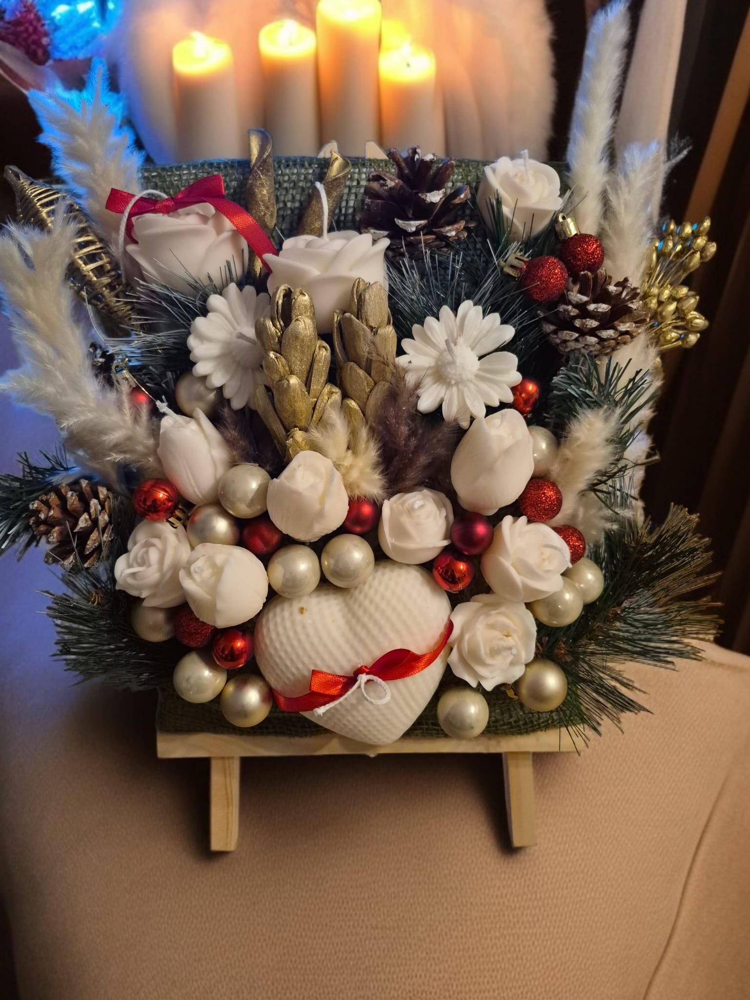 Decorative holiday floral arrangement on a small easel with white roses, berries, pinecones, and faux foliage, with warm glowing candles in the background.