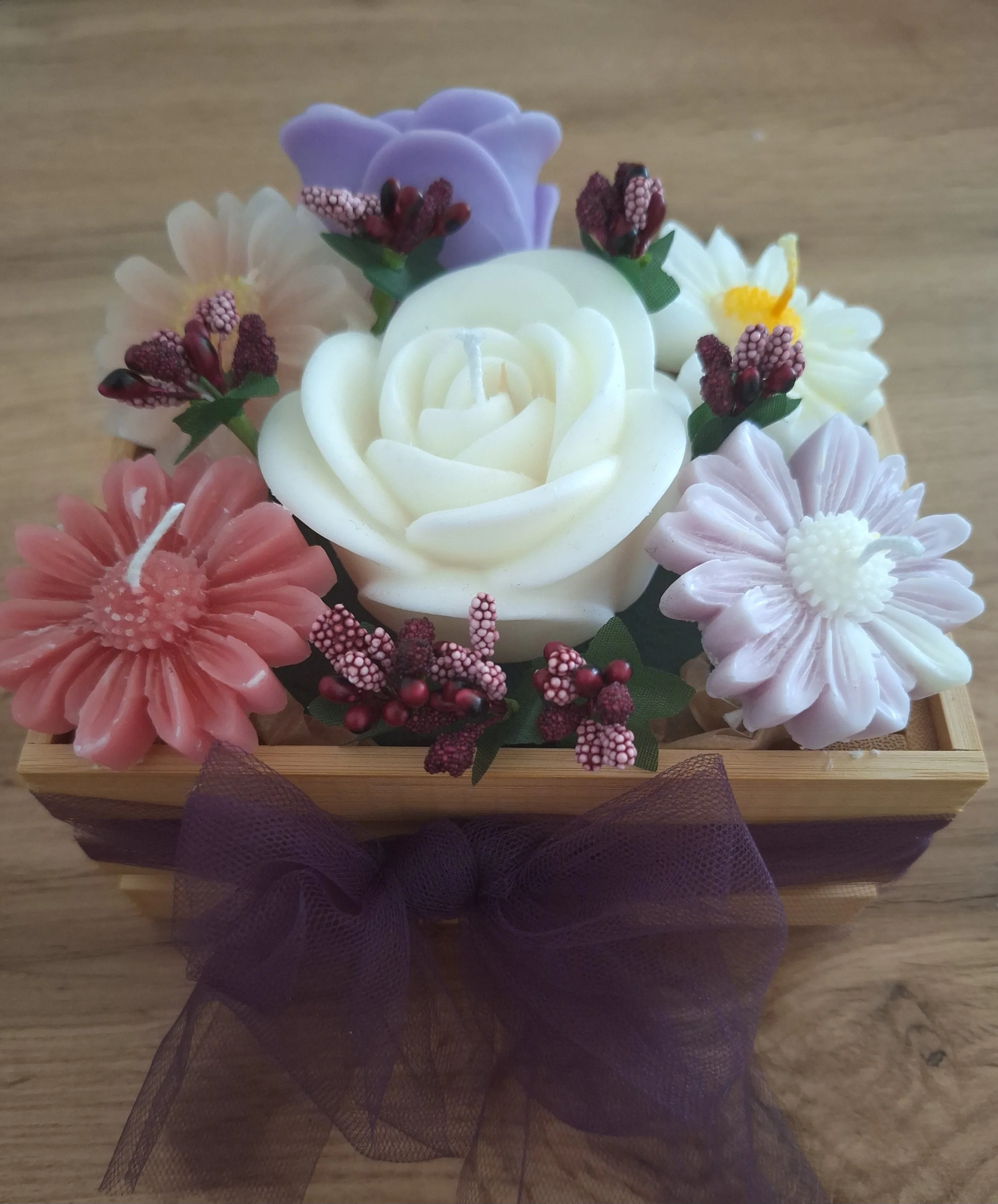 Kunstvolle Kerzen in Blumenform in einem kleinen Holzkörbchen, dekoriert mit einem violetten Schleifenband.