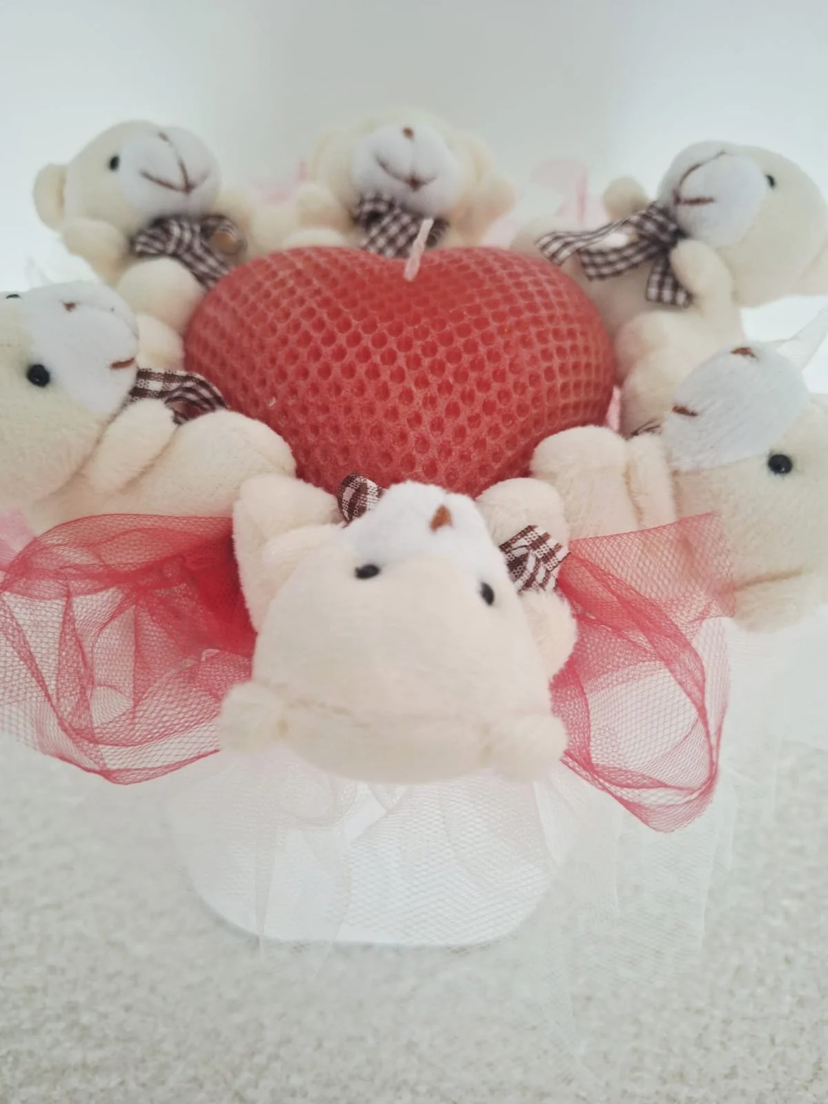 A collection of small white plush lamb toys with black eyes and brown stitched noses, each wearing a plaid bowtie, arranged around a red honeycomb-patterned heart-shaped item, with pink tulle fabric underneath.