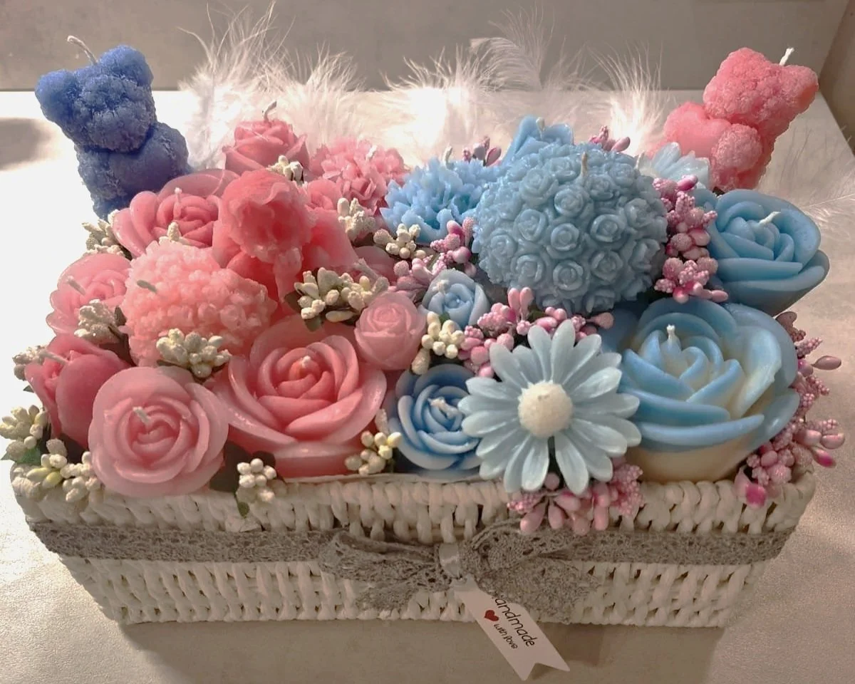 A decorative basket filled with pastel-colored artificial flowers including pink roses, blue roses, a white daisy, and various small flowers. There are also plush teddy bear shaped candles in blue and pink on top of the arrangement.