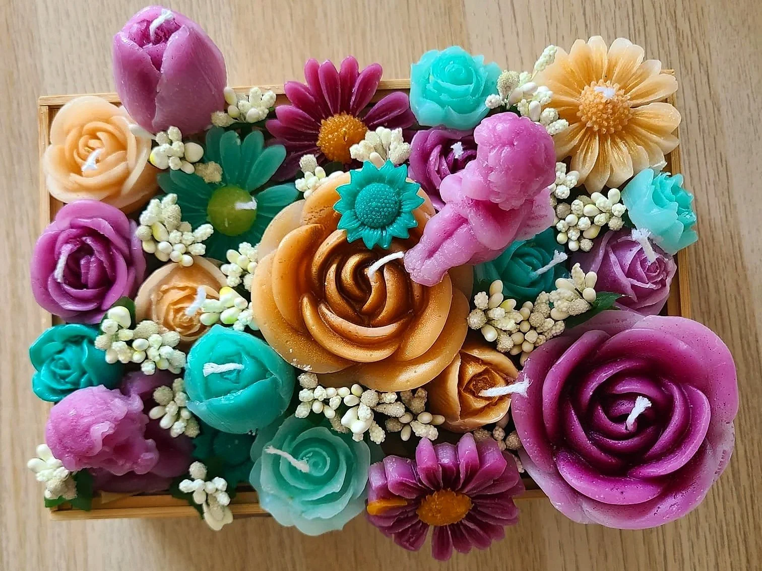Colorful assortment of flower-shaped candles in pink, purple, teal, peach, and yellow, arranged with small white faux flowers in a wooden box.
