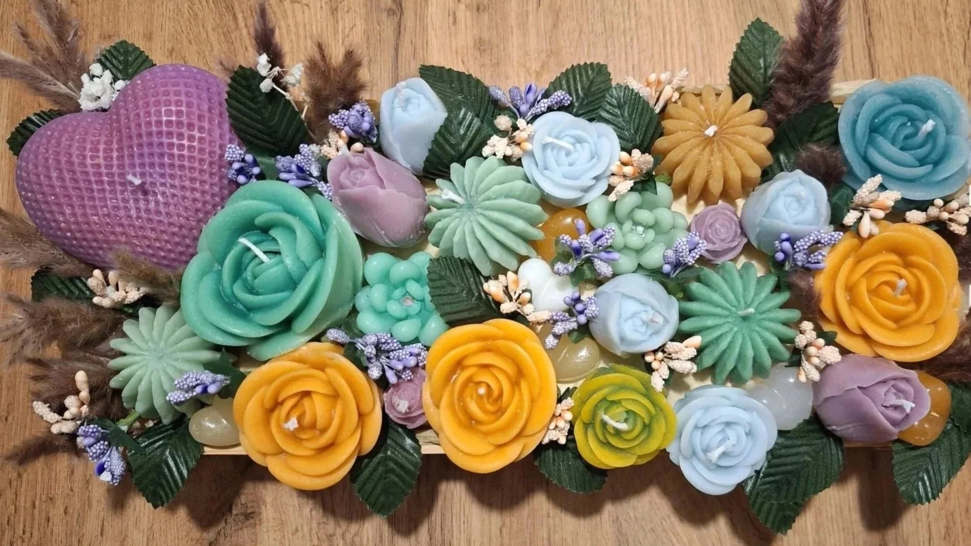 Colorful arrangement of candles shaped like roses, flowers, and a heart, with artificial leaves and small decorative flowers on a wooden surface.