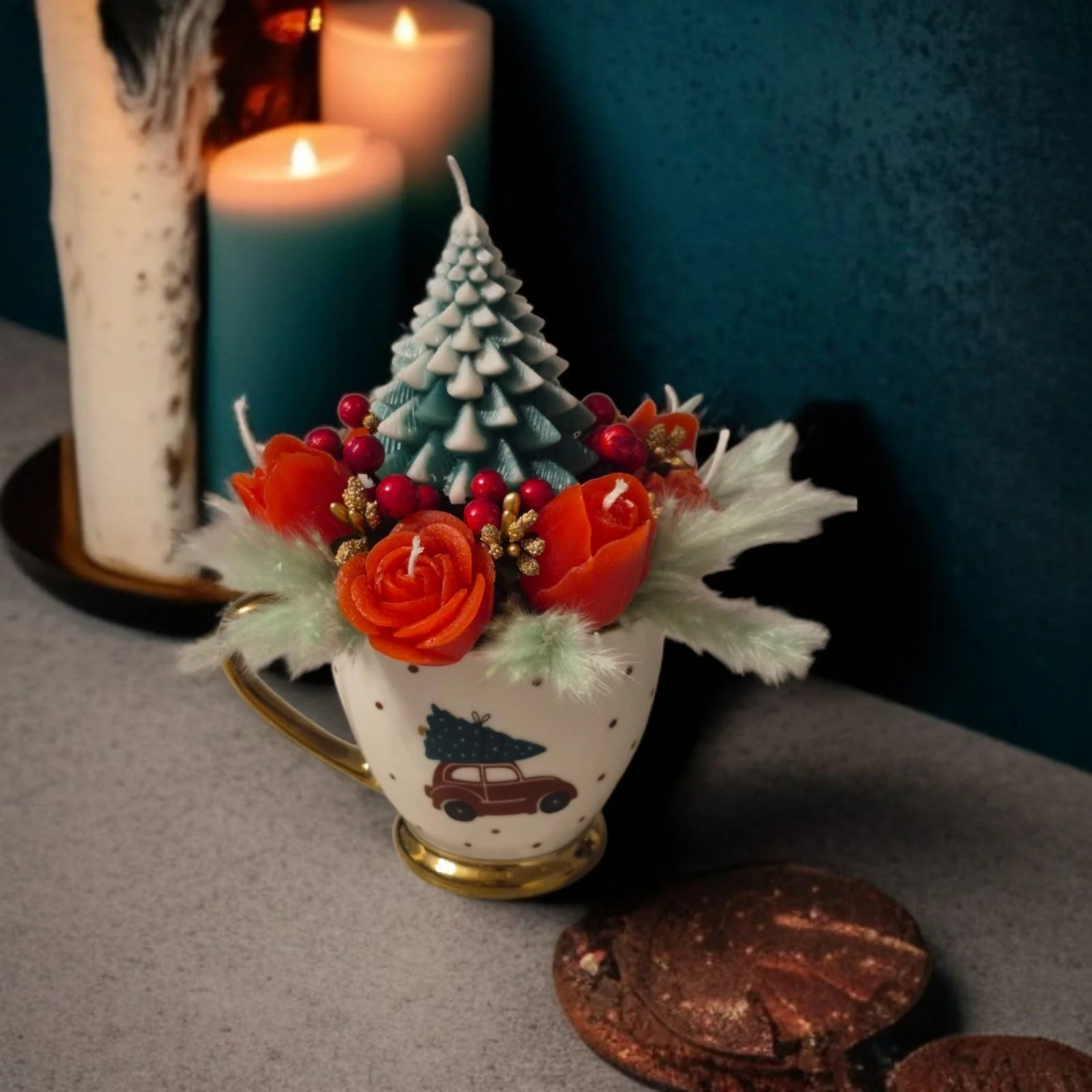 A decorated Christmas centerpiece with orange roses, red berries, a small blue-green Christmas tree, and gold accents in a white mug with a car and Christmas tree illustration. In the background, two lit candles, one blue and one white, on a black tray. A brown decorative object is in the foreground.