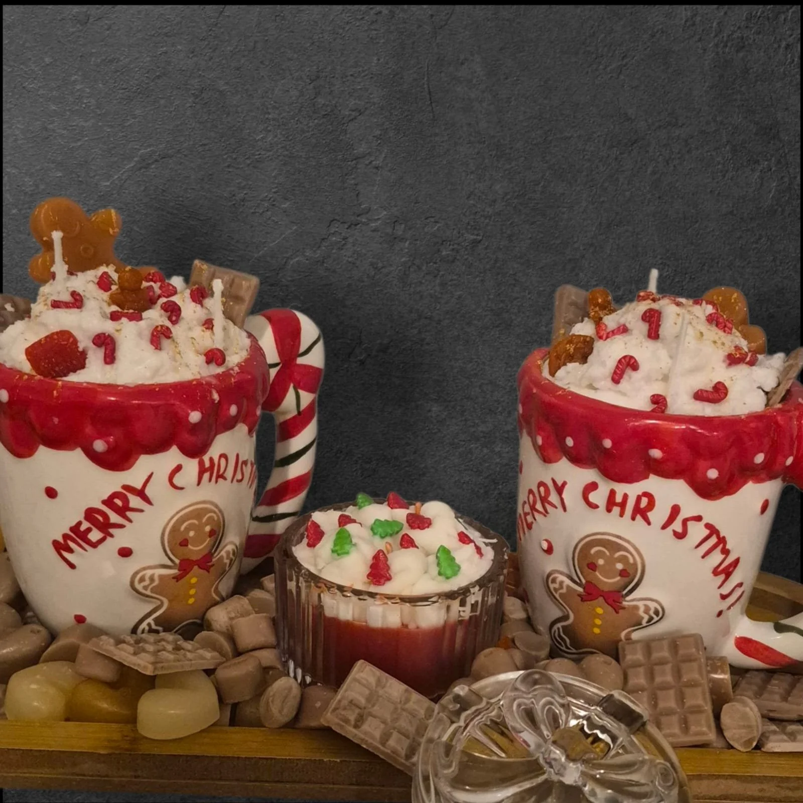 Two holiday-themed mugs with 'Merry Christmas' and gingerbread man designs, filled with whipped cream and decorated with candies and chocolates. A small jar of whipped cream with holiday sprinkles and an assortment of chocolates and candies surround the mugs, all set on a wooden tray against a dark background.