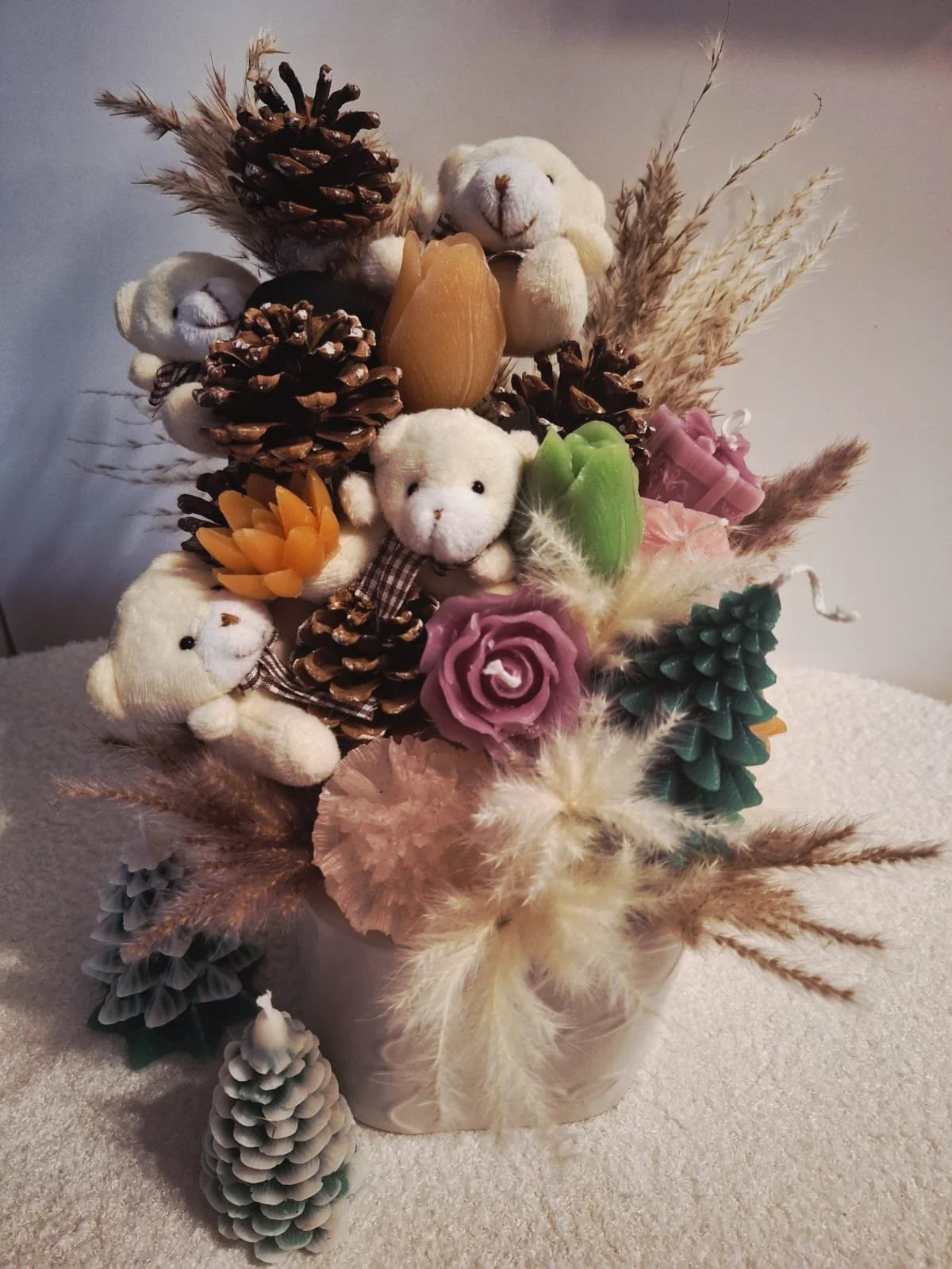 Decorative arrangement of small plush teddy bears, pinecones, candles, artificial flowers, and dried grasses in a white vase.