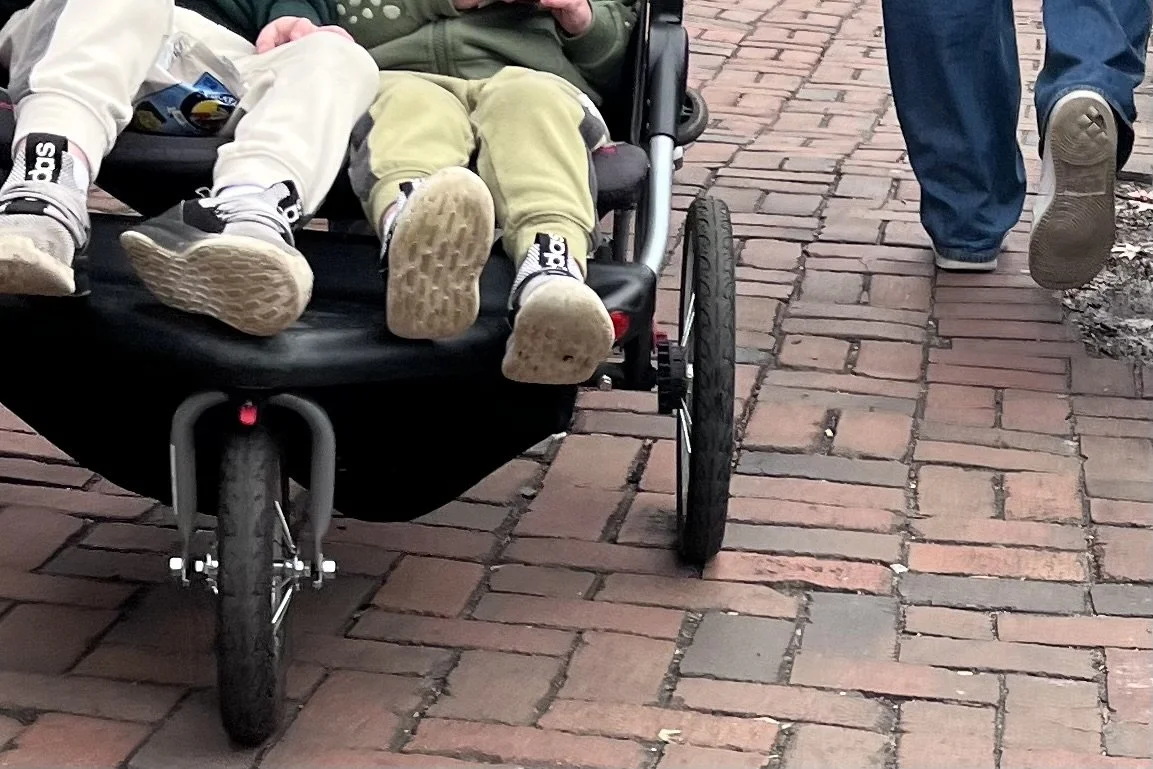 Stroller wheels and pedestrians moving along a narrow brick sidewalk in a busy historic district.