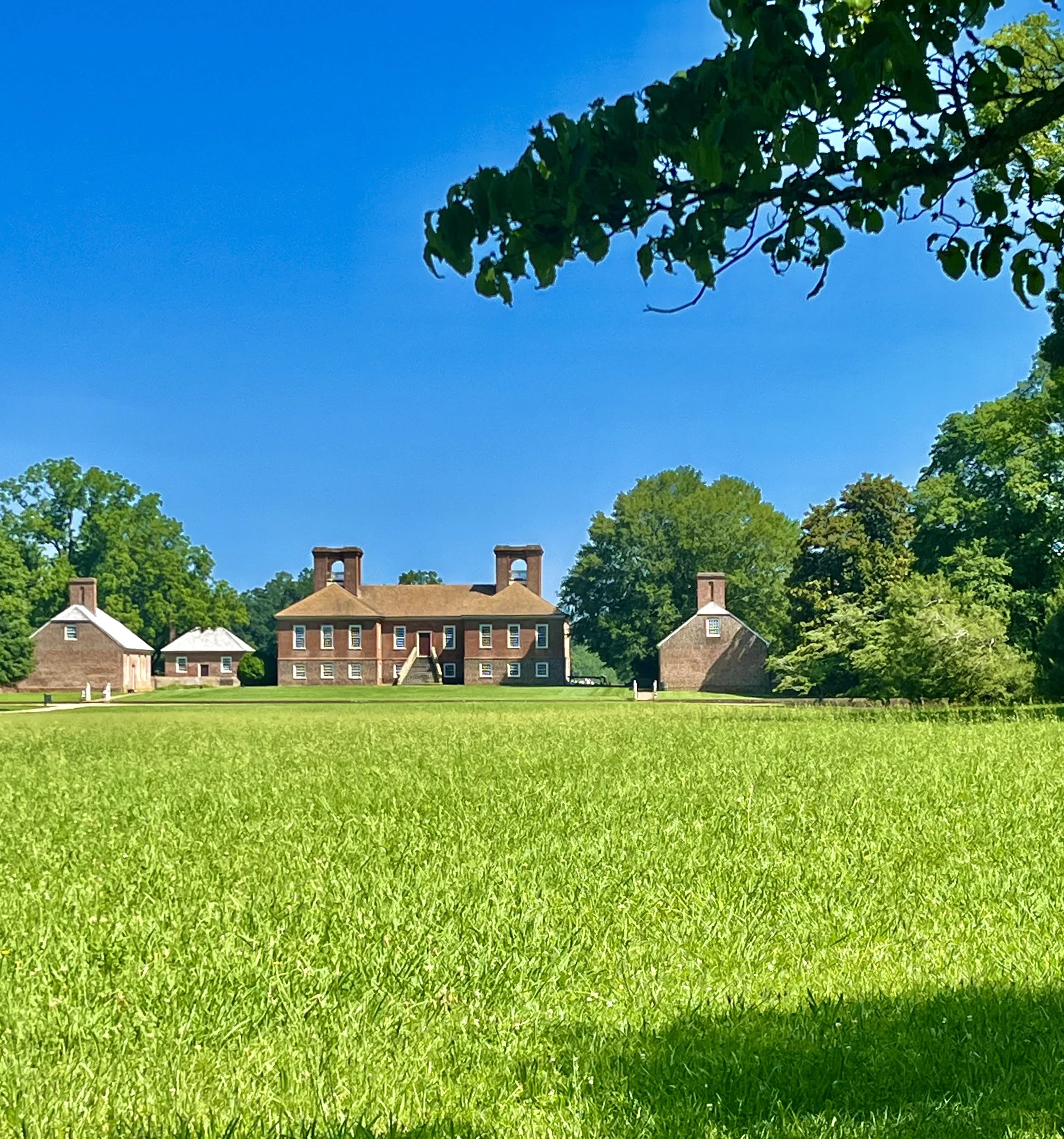 Wide green lawn leading to the red-brick buildings of Stratford Hall, framed by mature trees under a clear blue sky in Westmoreland County, Virginia.