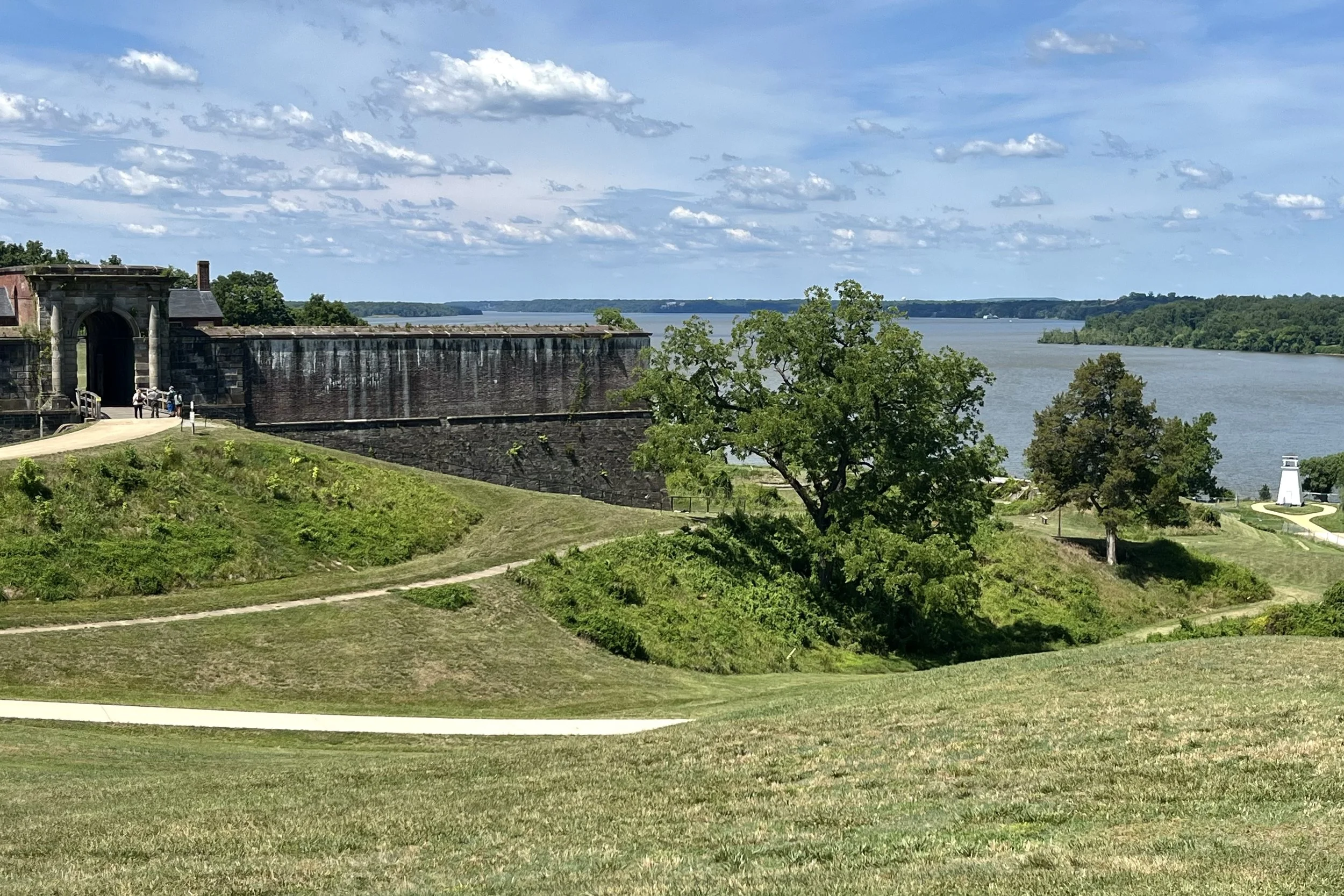View from Fort Washington Park overlooking the Potomac River, with historic brick fort walls, grassy earthworks, walking paths, and river scenery under a blue sky with scattered clouds.