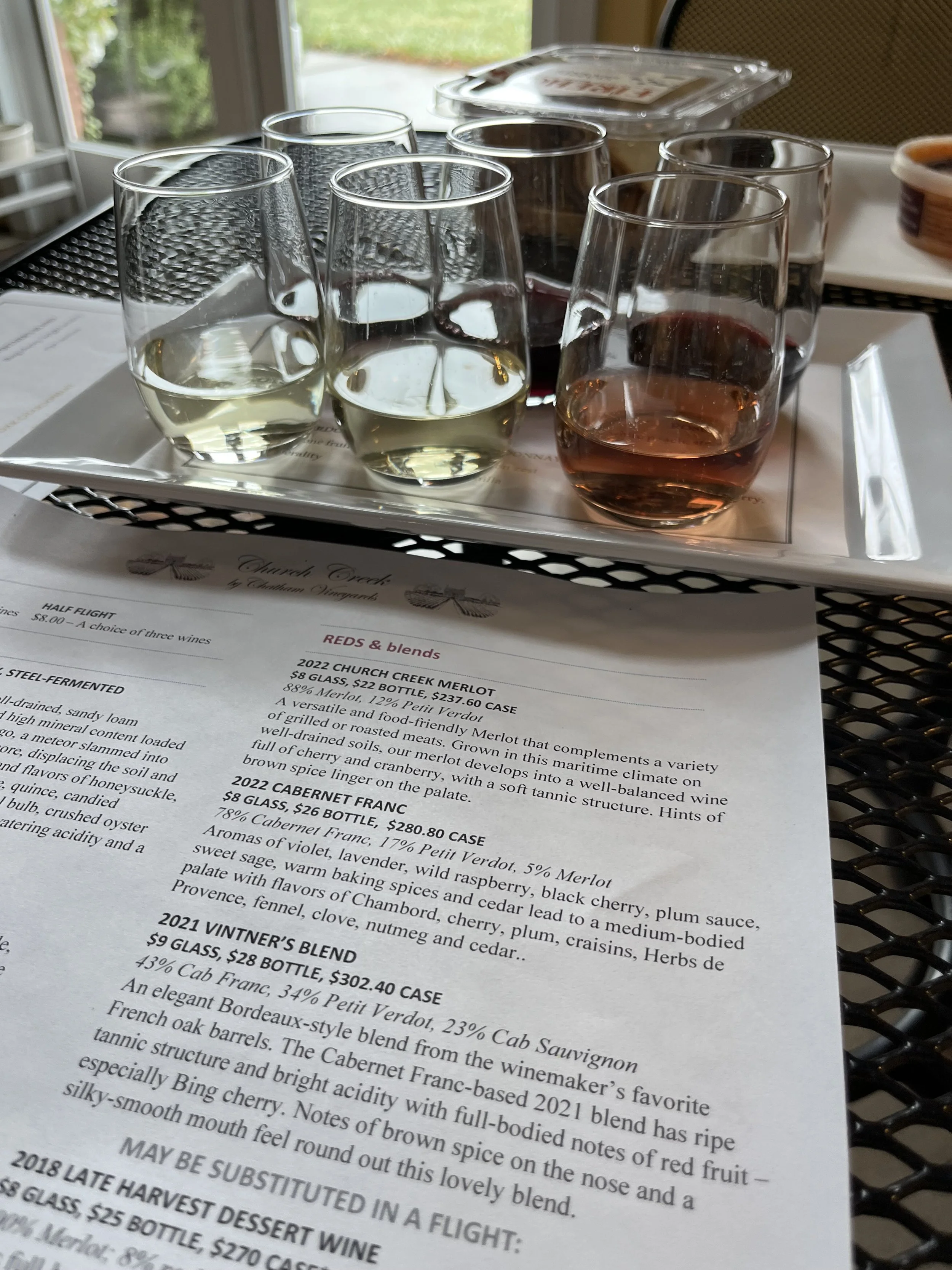 Six wine glasses arranged on a tasting tray, set on a table with a vineyard wine list beneath.