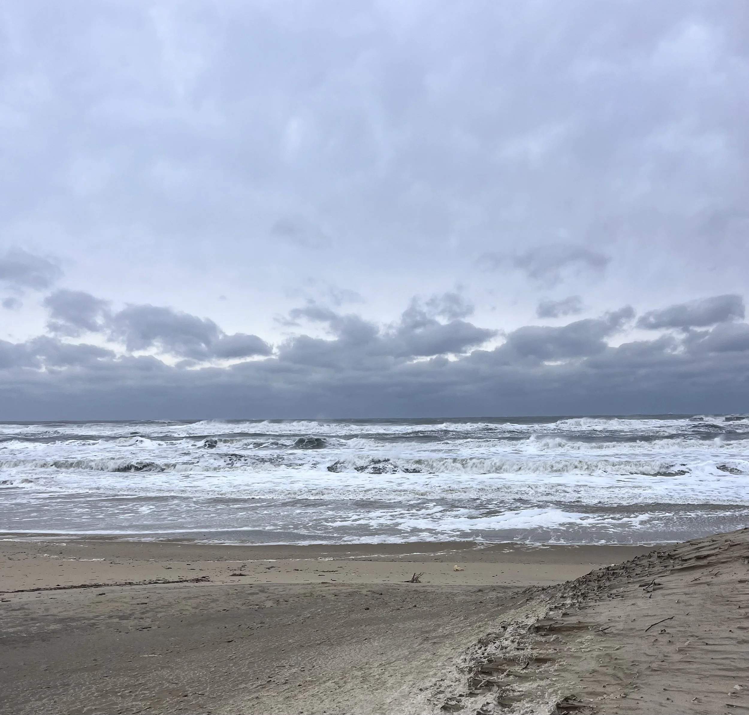 Wide view of a winter beach in the Outer Banks with rough ocean waves, wet sand, and layered gray clouds stretching across the horizon.