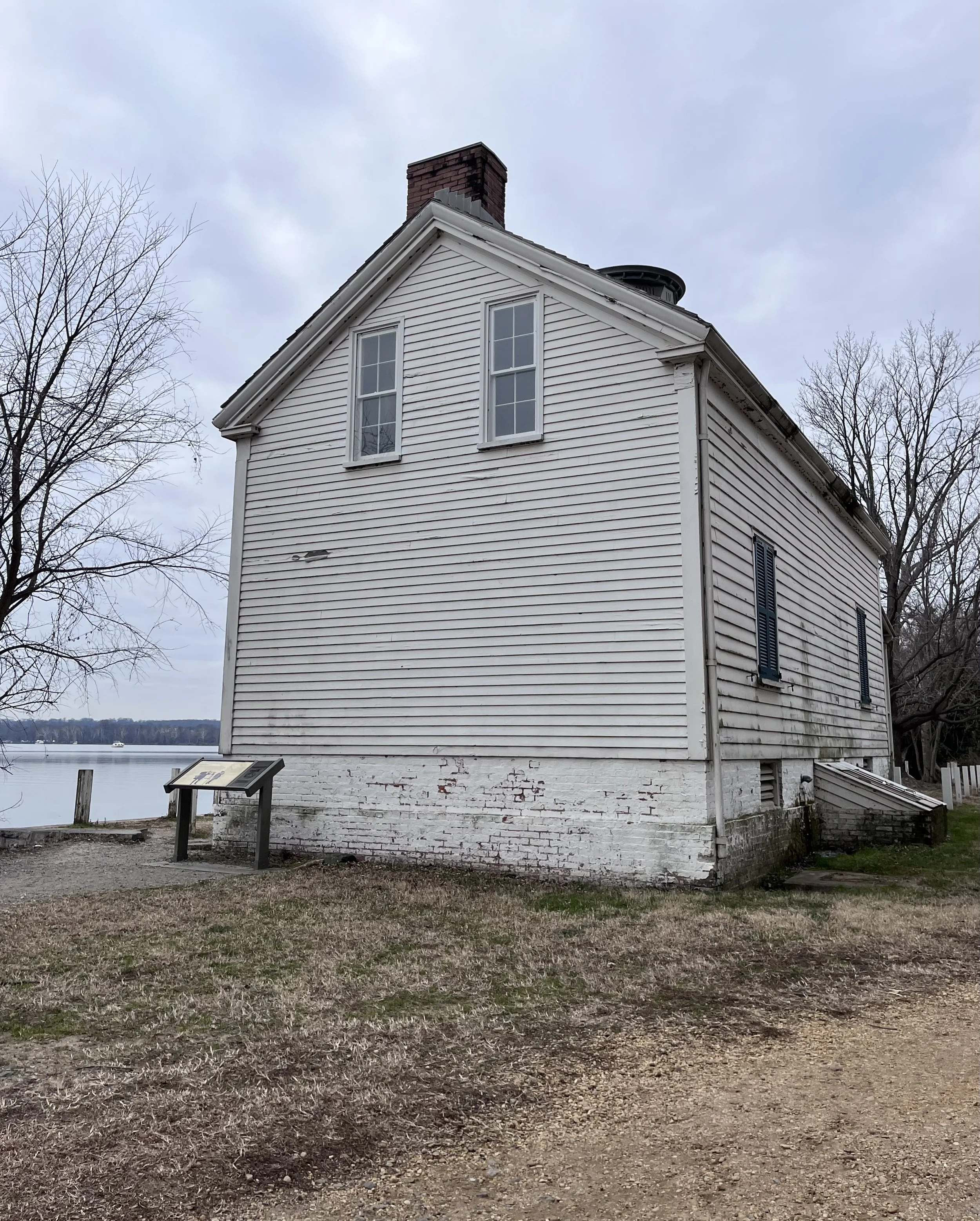 The historic white wooden keeper’s house at Jones Point Lighthouse viewed from the side, with the Potomac River visible behind it on an overcast day.
