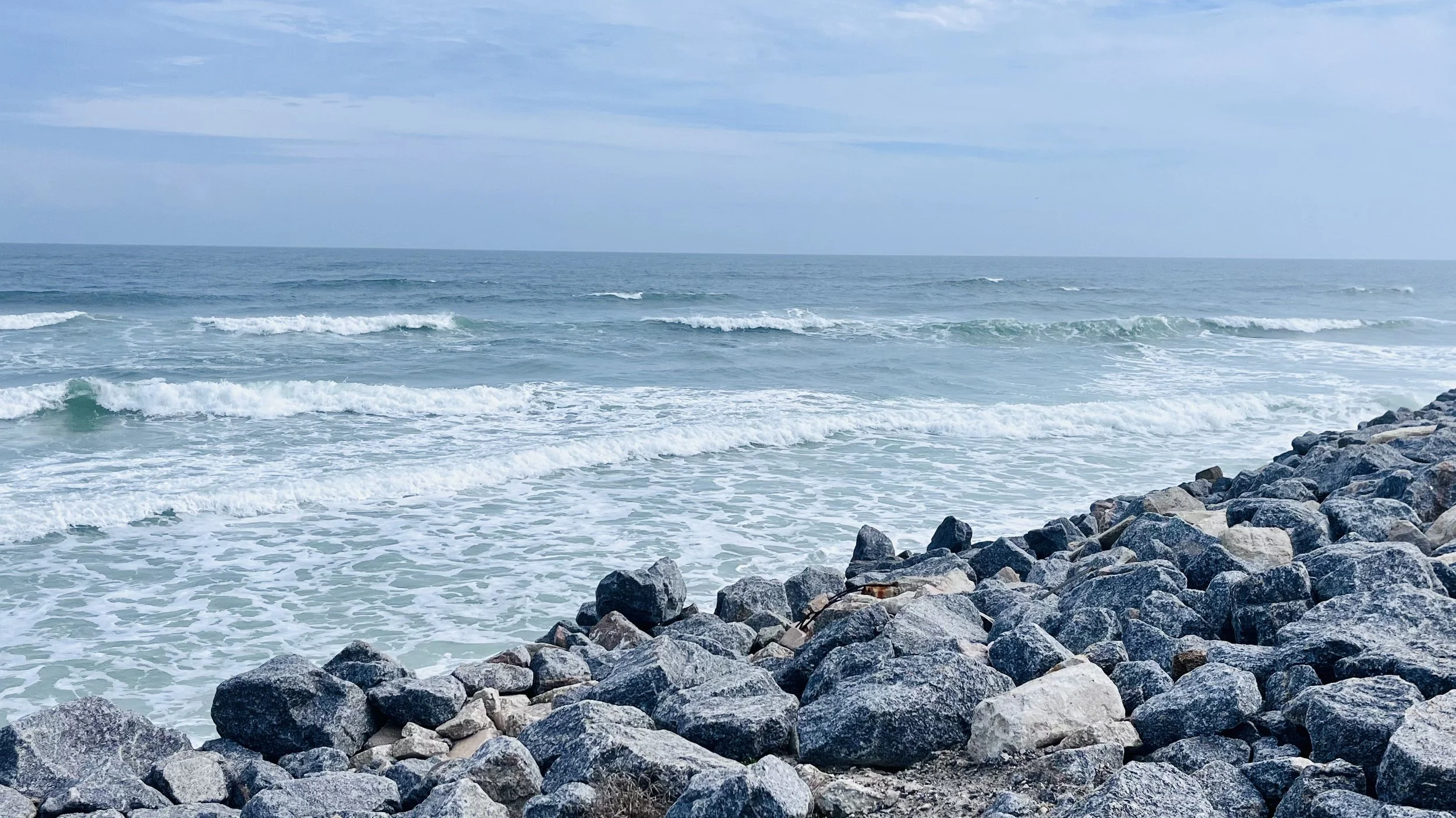 Waves roll toward a rocky shoreline under a pale blue sky, the ocean stretching calmly to the horizon.