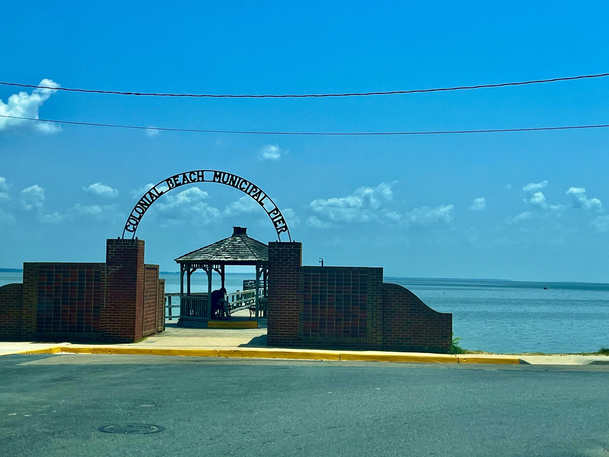 Brick entrance arch reading “Colonial Beach Municipal Pier,” leading to a wooden gazebo on the pier overlooking calm Potomac River waters under a lightly clouded blue sky.