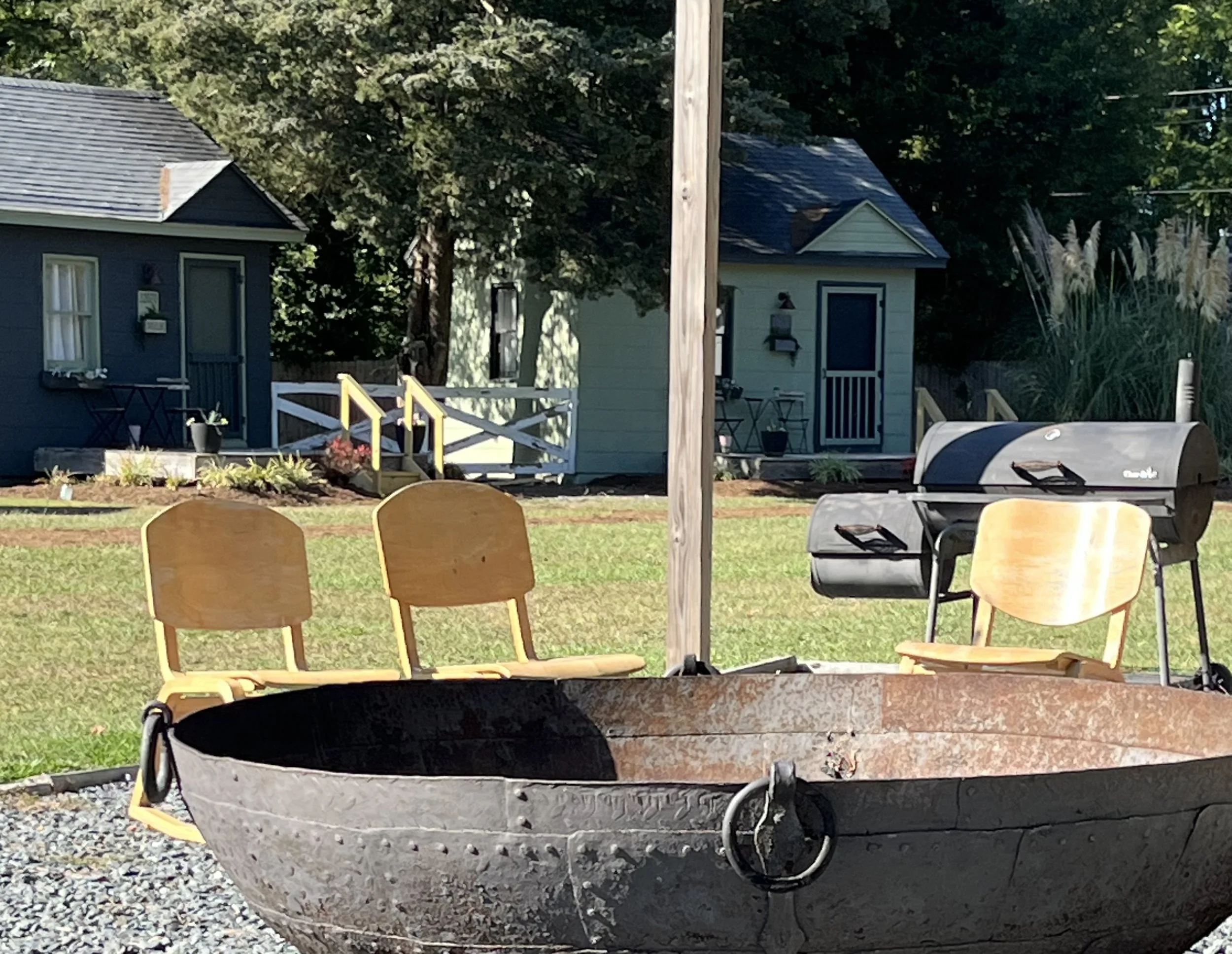 Large metal fire pit with chairs around it, set on a grassy area near small cabins.