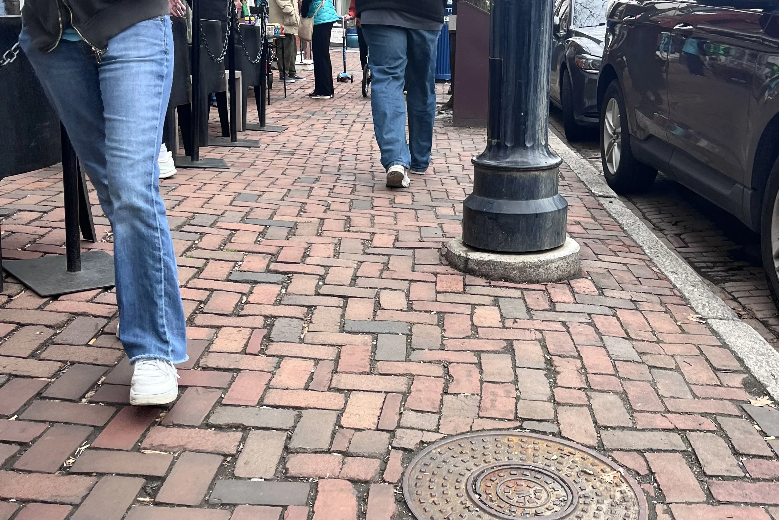 Pedestrians walking along a narrow brick sidewalk beside parked cars in a busy historic district.