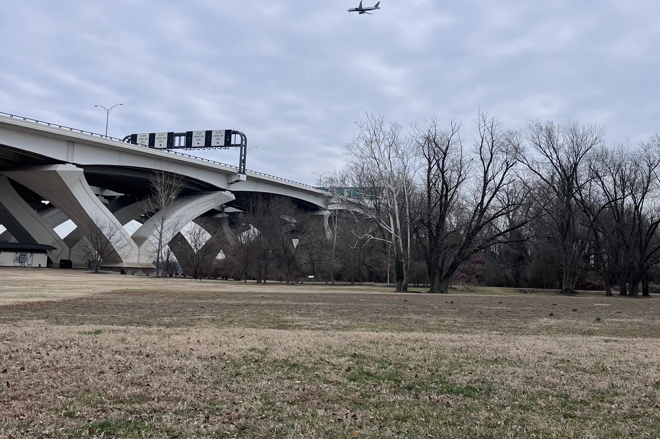 Wide grassy field at Jones Point Park beside the large bridge structure, with open space, trees, and an airplane overhead.