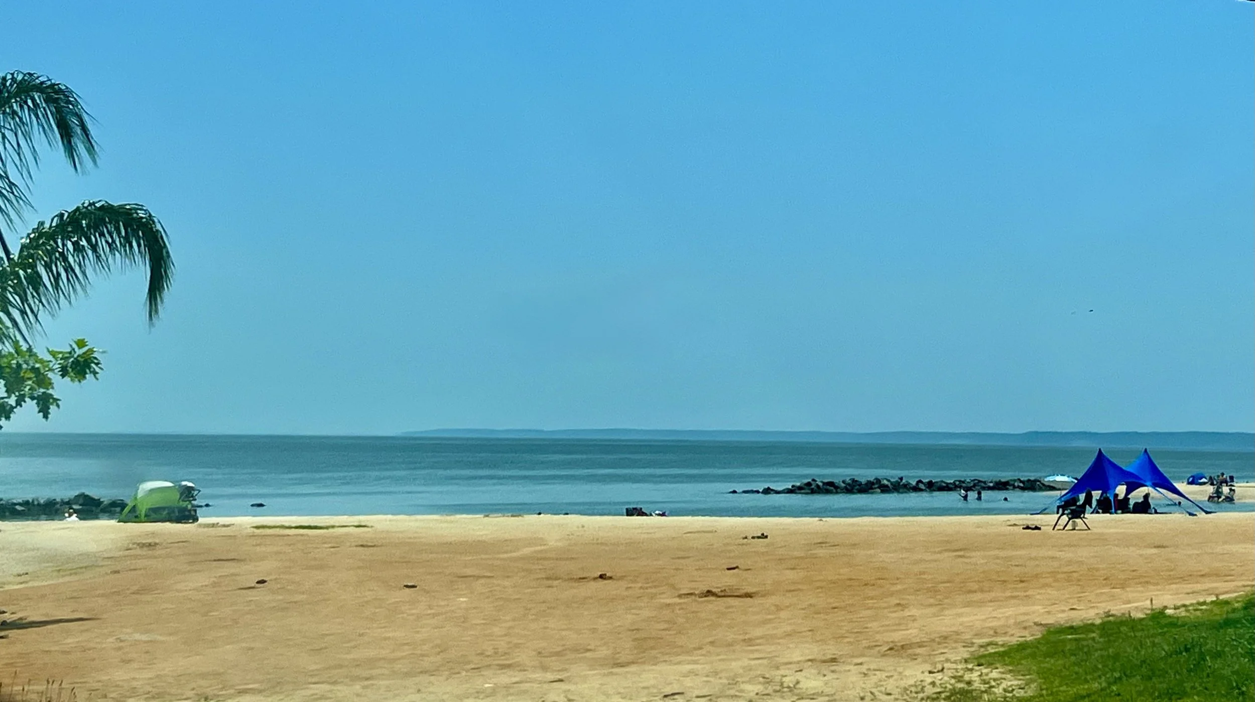 Wide sandy beach at Colonial Beach, Virginia, with calm Potomac River waters, a clear blue sky, small rock jetty in the distance, and a few beach tents along the shoreline.