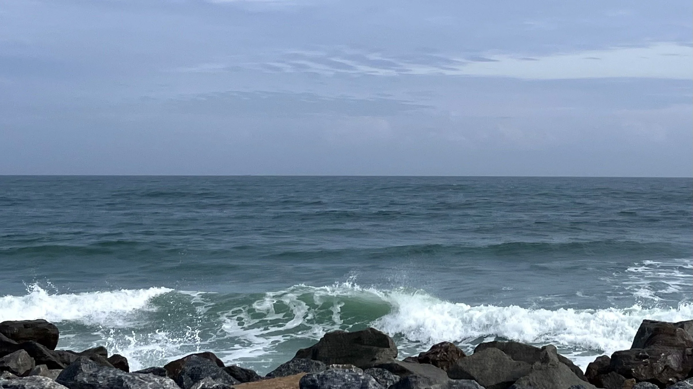 Ocean waves rolling in at Matanzas Inlet, with dark rocks in the foreground and a cloudy sky above