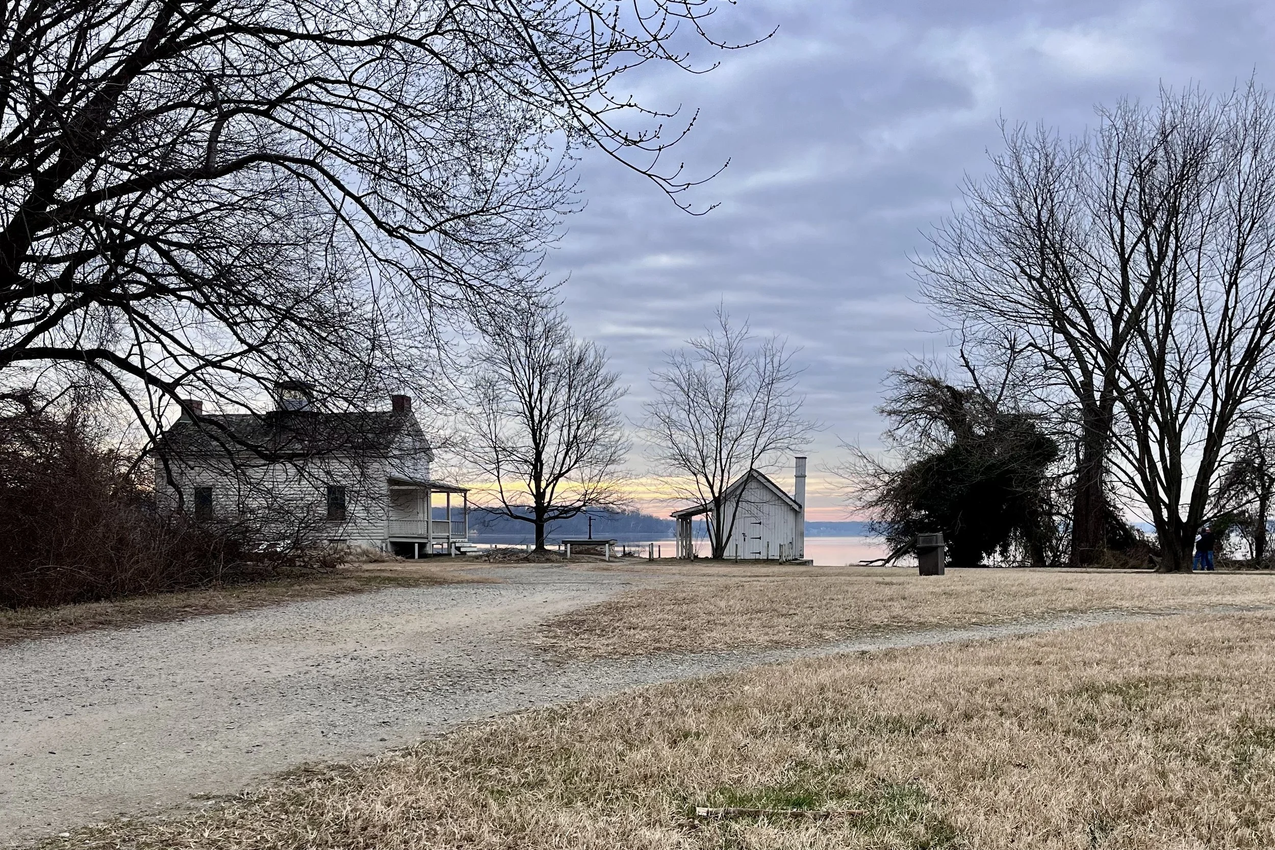 Curved gravel path leading toward the Jones Point lighthouse area at sunset, with open grass, bare trees, and water visible in the background.