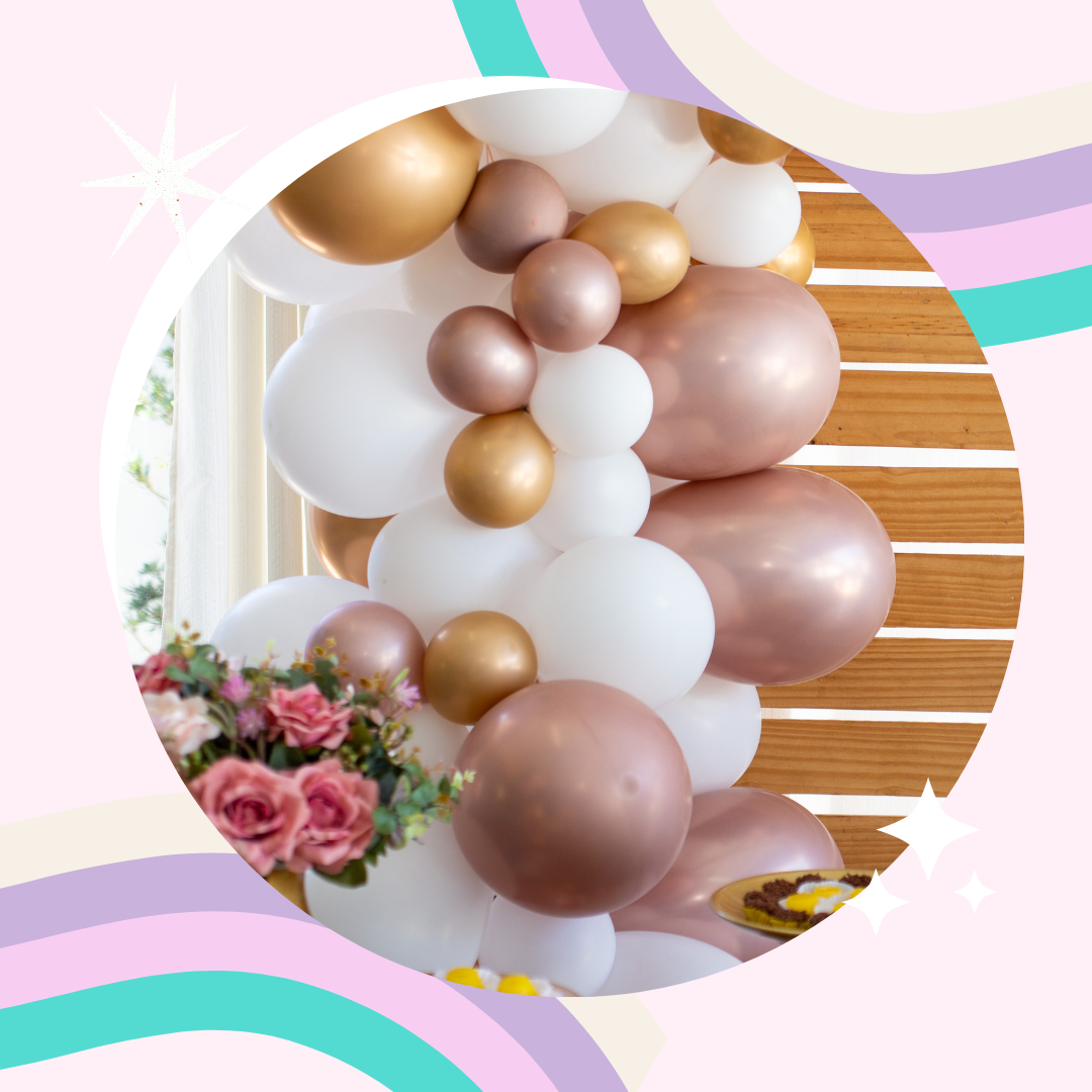 A decorative balloon bouquet consisting of white, gold, bronze, and rose gold balloons with pink flowers in the foreground, set against a wooden background and bright window.