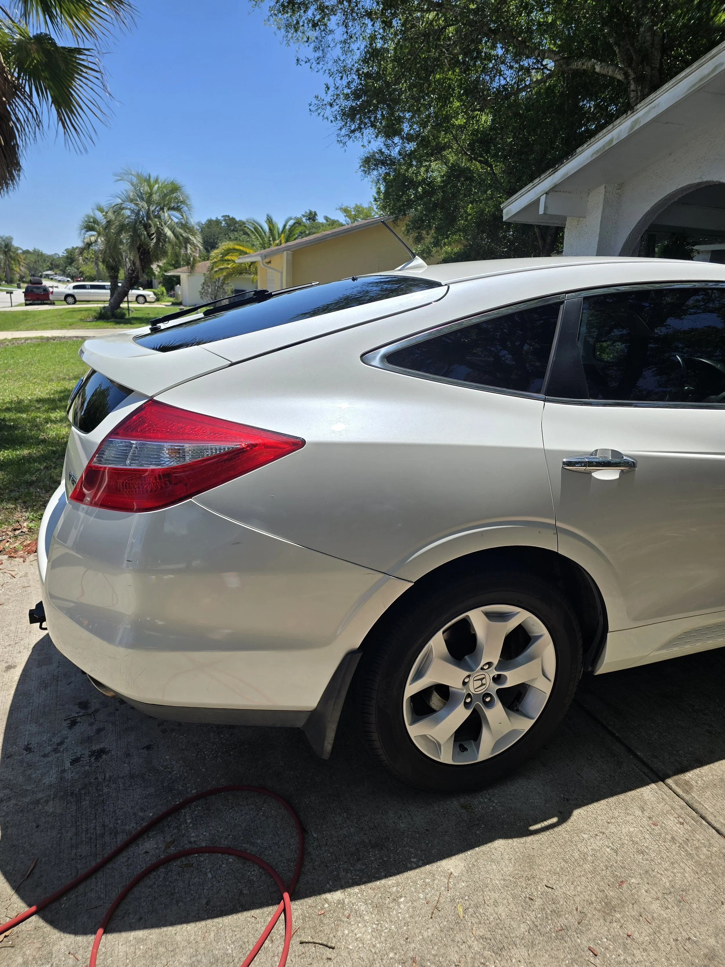 White Honda sedan parked on a driveway with a garden hose on the ground nearby, trees, and houses under a clear blue sky.