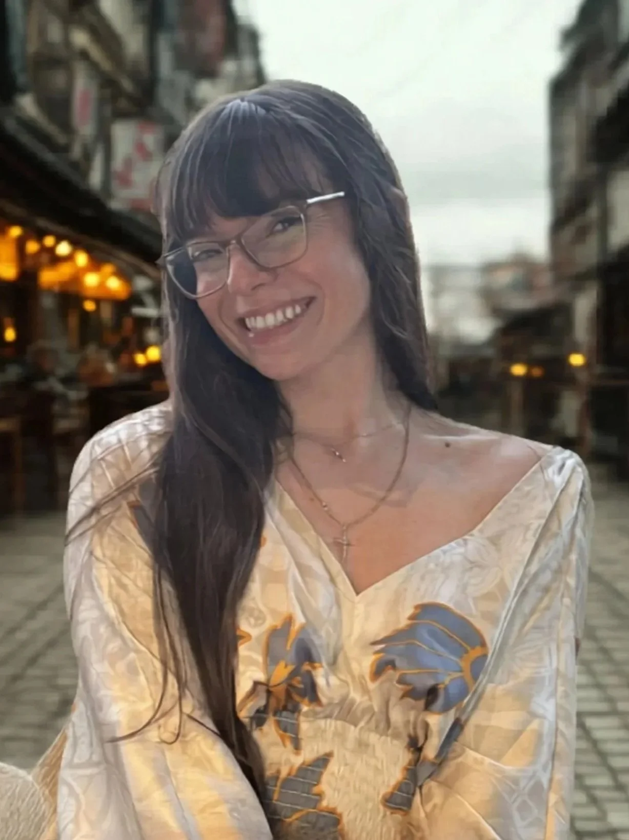 A smiling woman with long dark hair, glasses, and layered necklaces, wearing a beige and yellow patterned top, in a warmly lit outdoor restaurant or café.
