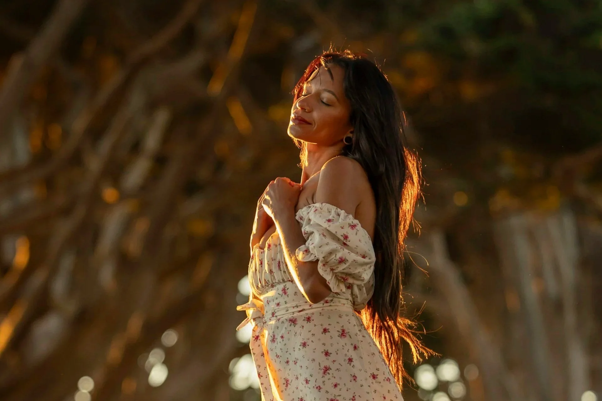 A woman with long dark hair is outdoors during sunset, wearing an off-shoulder floral dress, with eyes closed and a gentle smile, embracing herself.