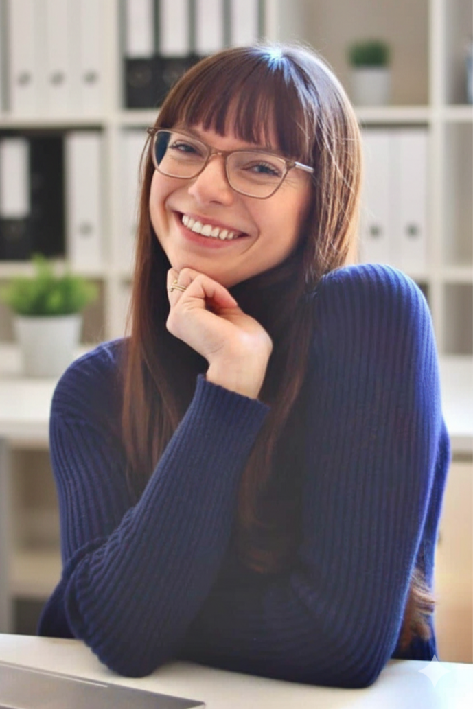 A woman with long brown hair, wearing glasses and a blue sweater, smiling with her chin resting on her hand in an office environment with bookshelves and a potted plant in the background.