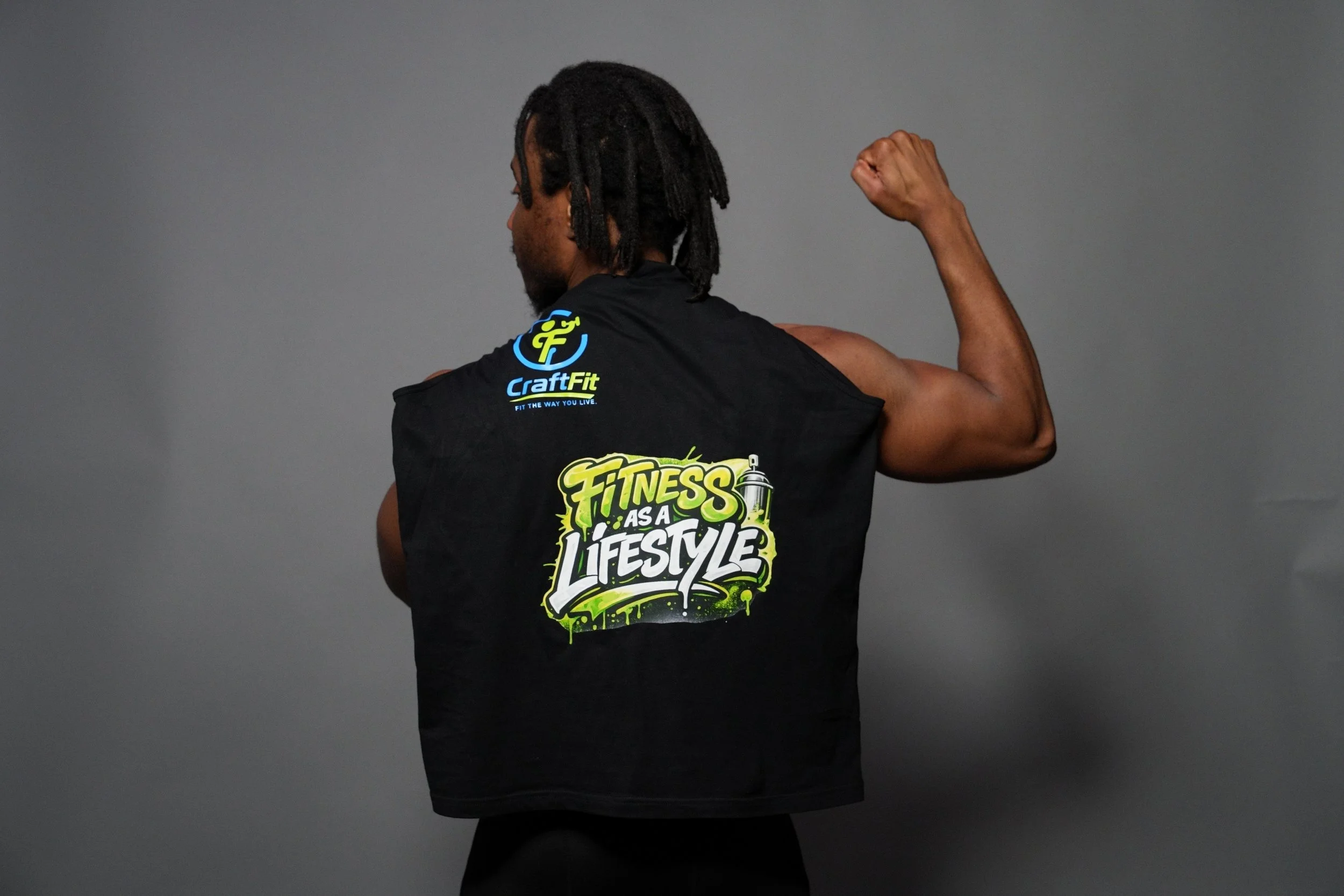 Back view of a person with dreadlocks flexing their right arm, wearing a black sleeveless shirt with colorful fitness-related logos and slogans, standing against a plain gray background.