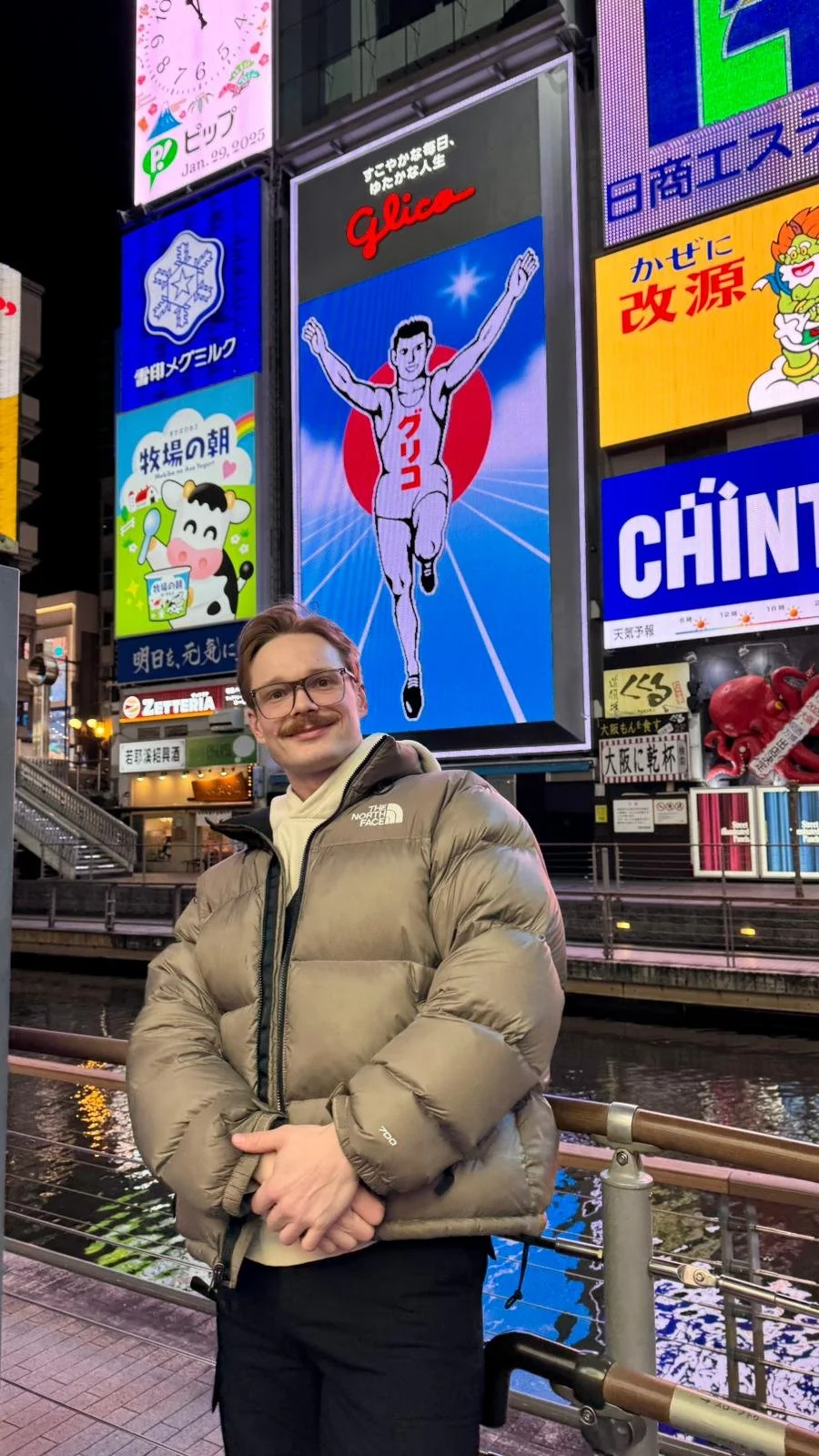A man with glasses and a beige puffy jacket standing in a city area at night with bright neon billboards in the background, including a large digital illustration of a running man with outstretched arms and the word 'Glico' in red.