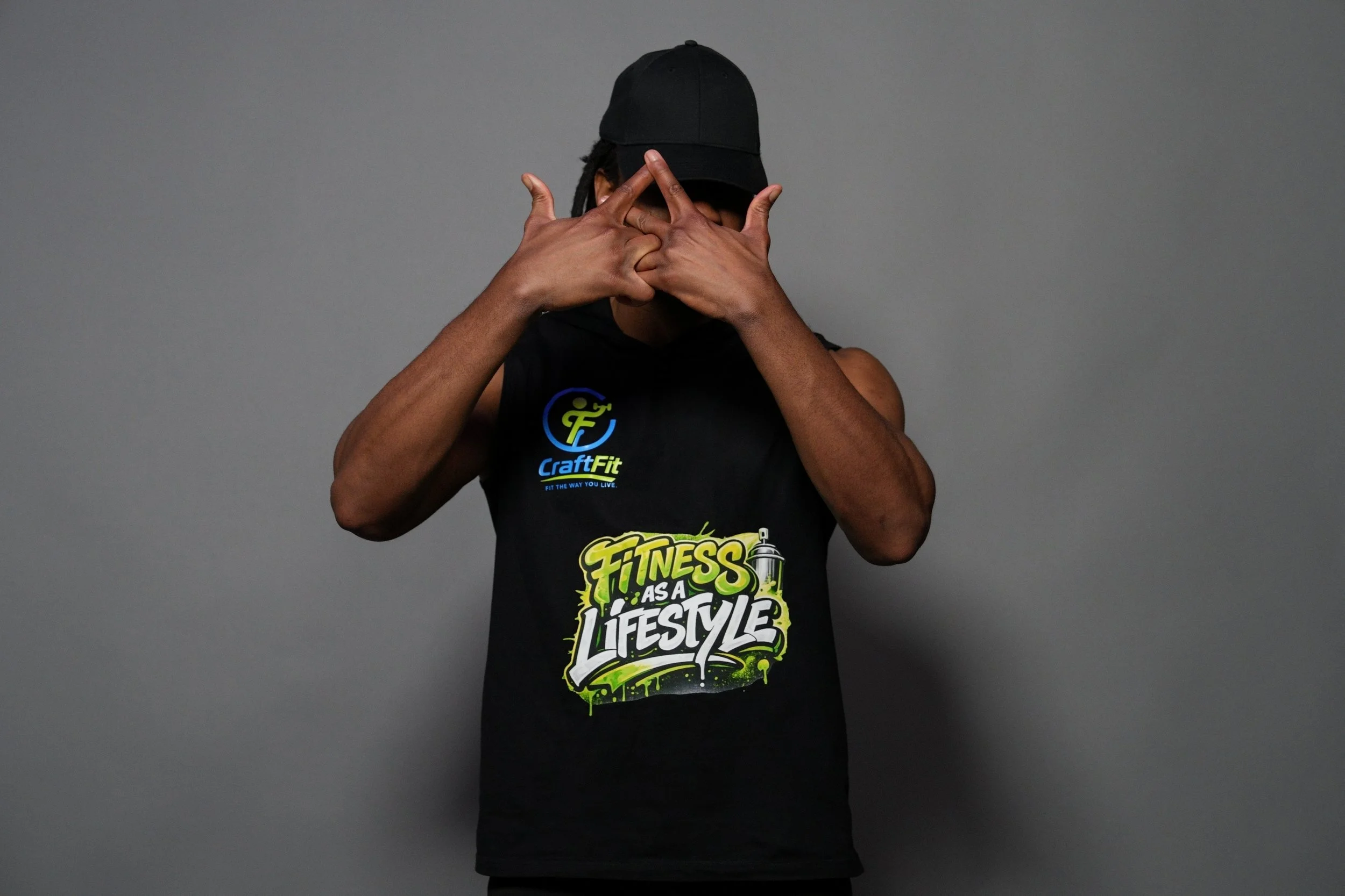 Person wearing a black Cap, covering face with hands, wearing a black sleeveless shirt with colorful fitness slogan, standing against plain gray background.