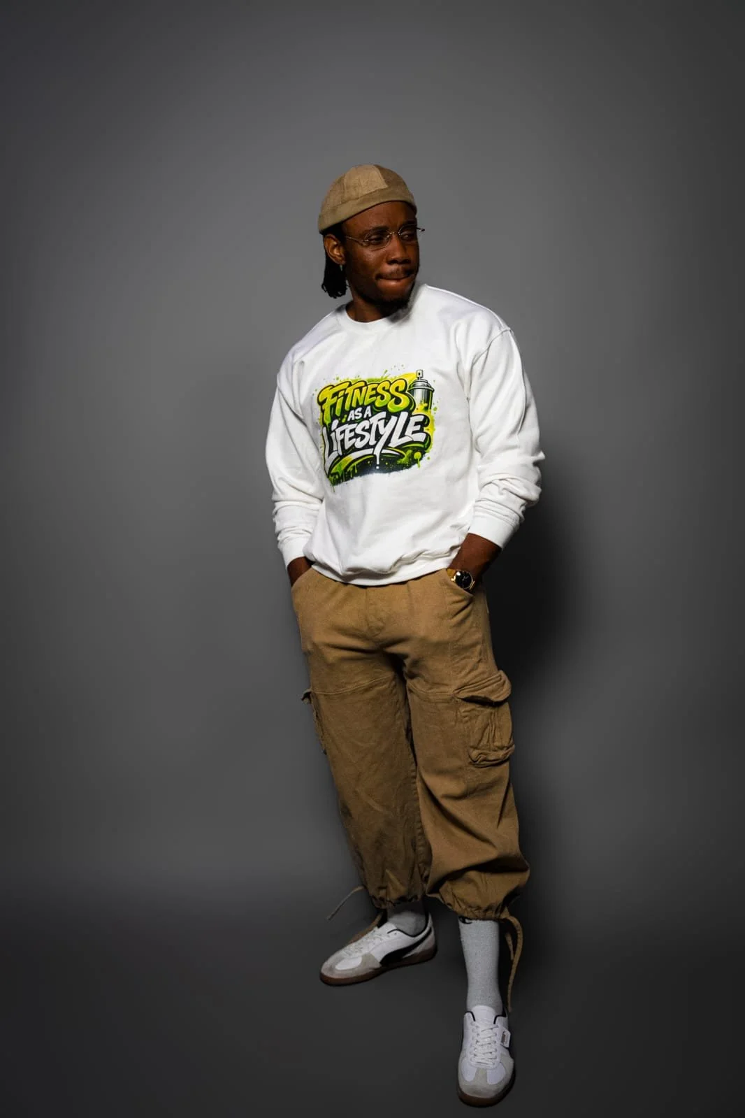 A young man standing against a gray background, wearing a white sweatshirt with green, yellow, and black graphic text that reads 'Fitness as a Lifestyle,' khaki cargo pants, white sneakers, gray socks, a tan cap, and glasses.