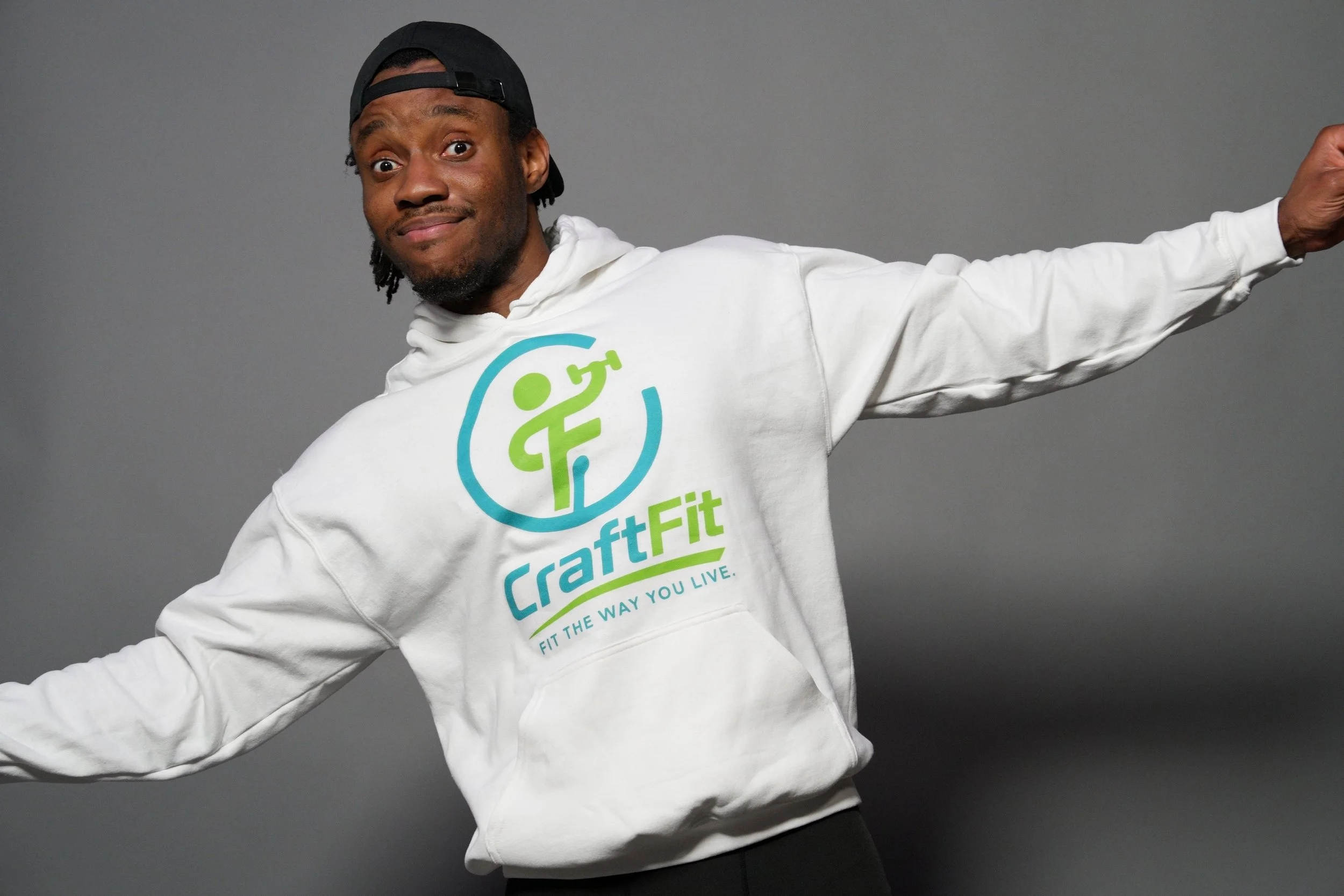 Person wearing a white hoodie with a logo, black cap worn backwards, arms outstretched, against a plain gray background.