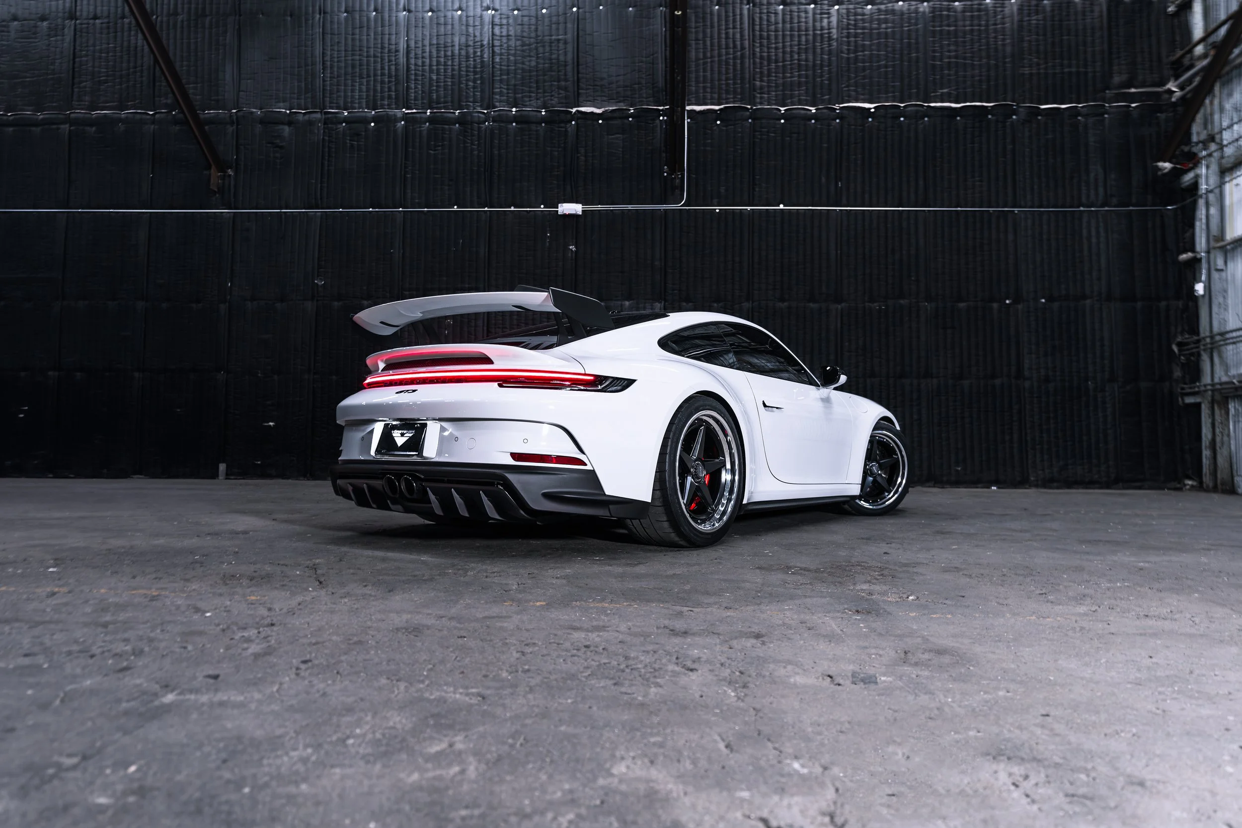 White sports car with a large rear wing and black rims parked in an indoor garage with dark walls.