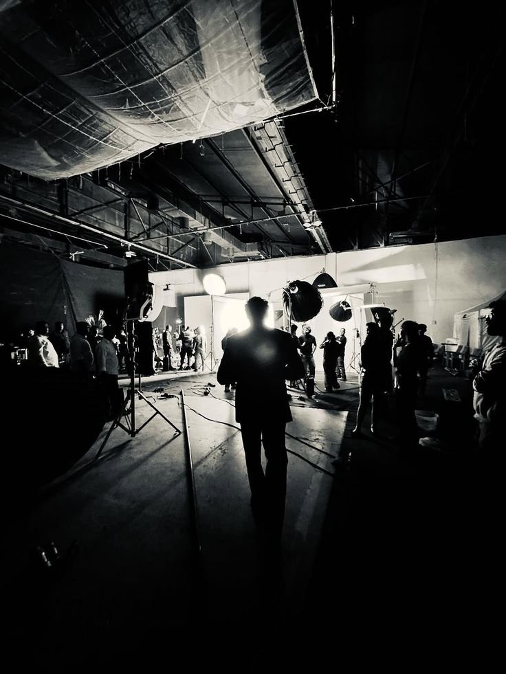 Behind-the-scenes view of a film or photography studio with crew members, lighting equipment, and backdrops, all in black and white.