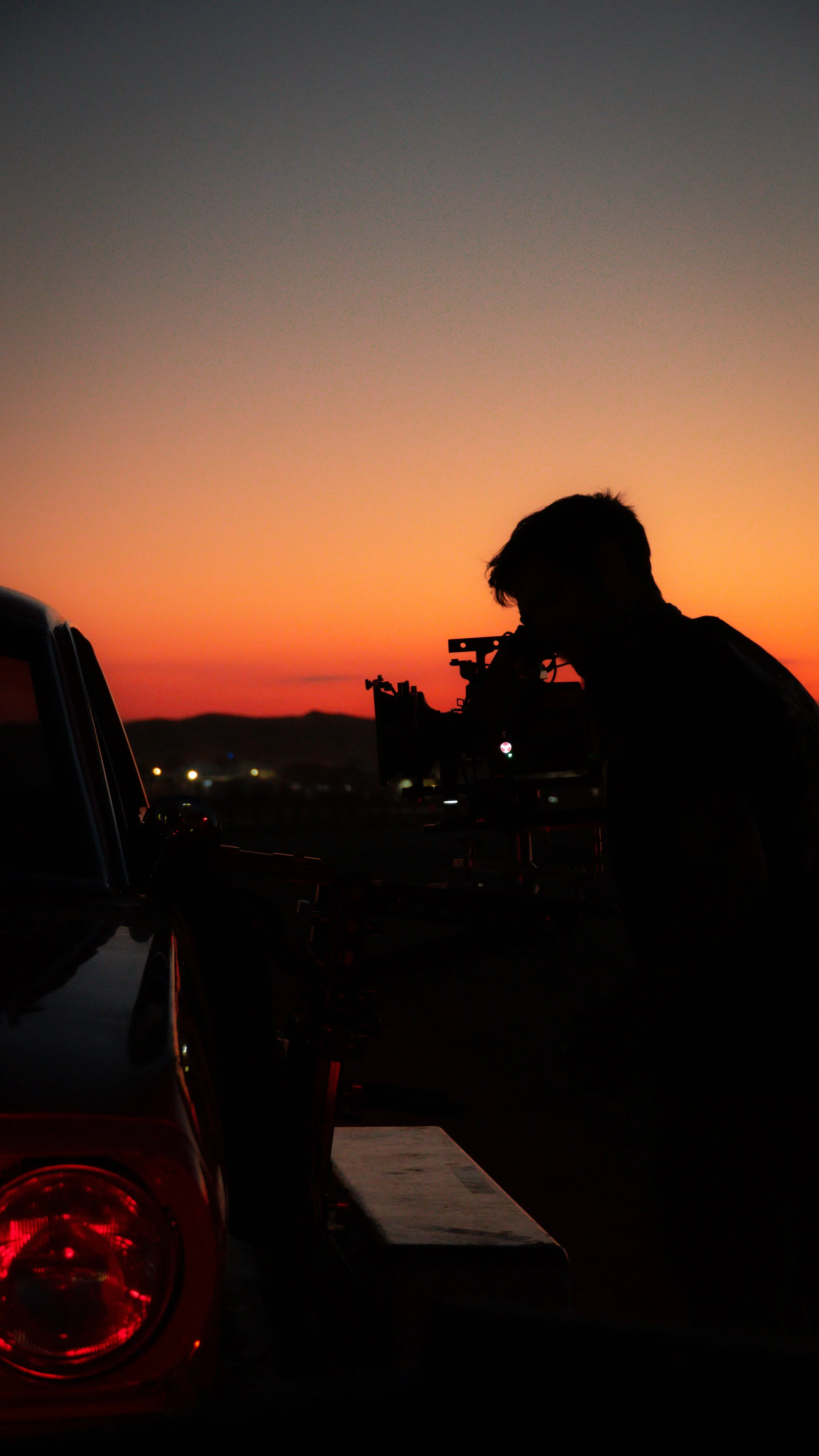 Silhouette of a person operating a camera on a tripod during sunset at dusk with a car and distant hills in the background.
