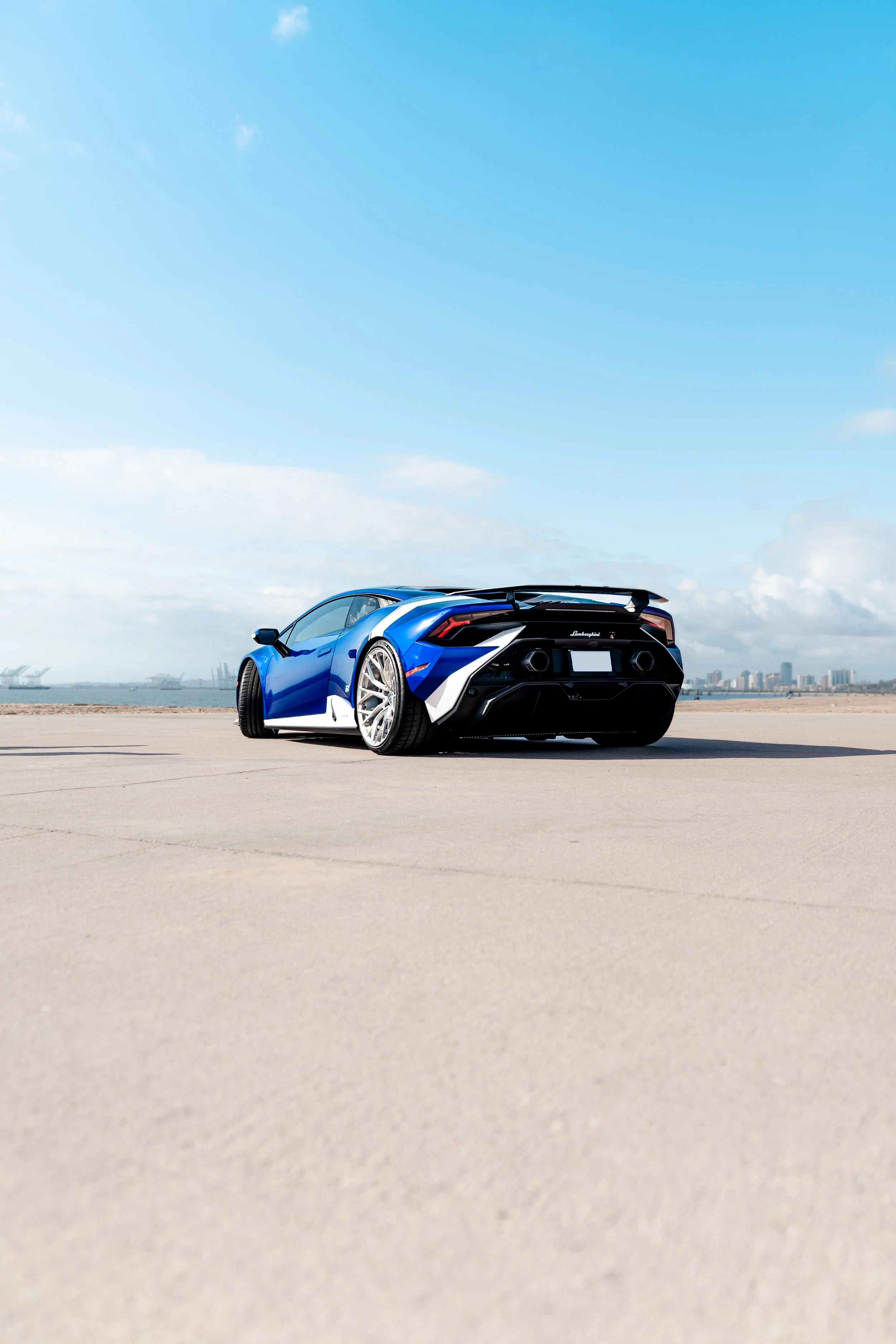 Blue Lamborghini sports car parked on a concrete surface with a city skyline and water in the background under a partly cloudy sky.