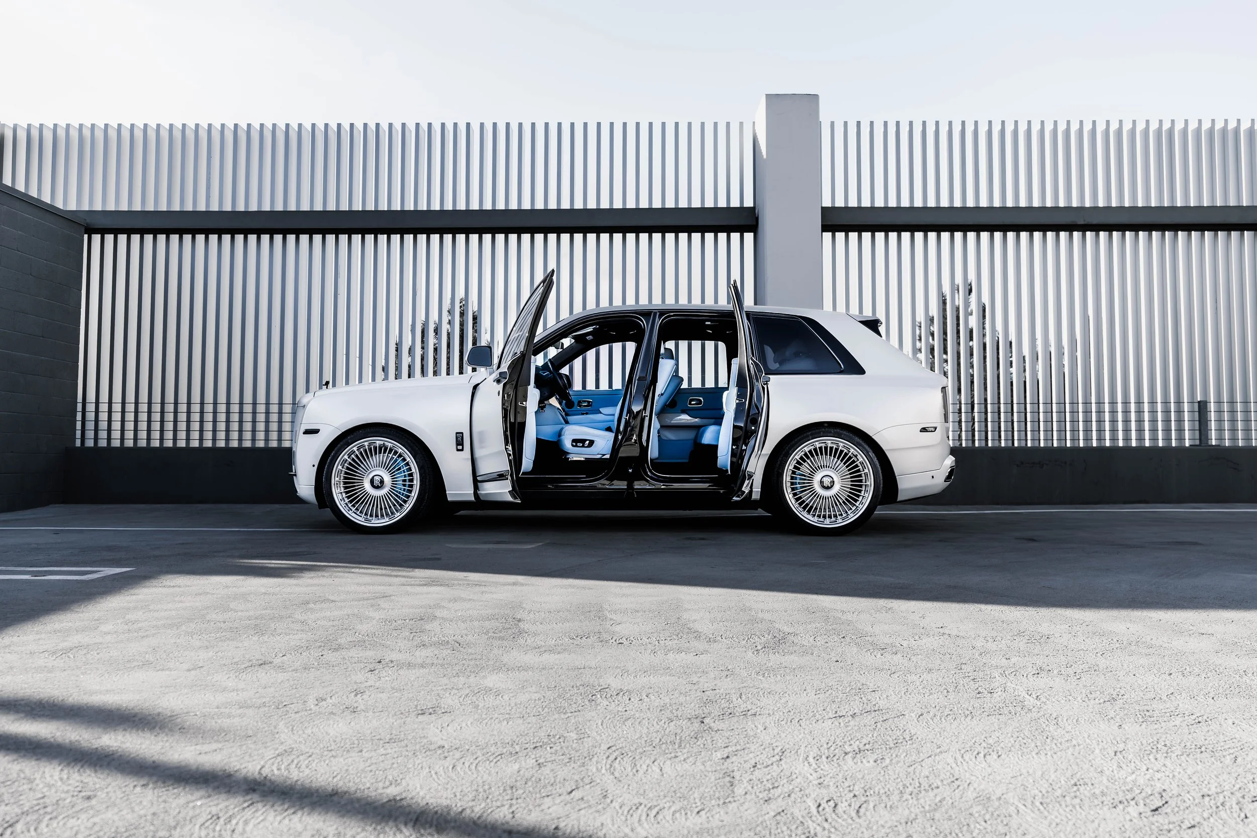 A white luxury car with all doors open, parked in a modern outdoor space with a geometric metal fence in the background.
