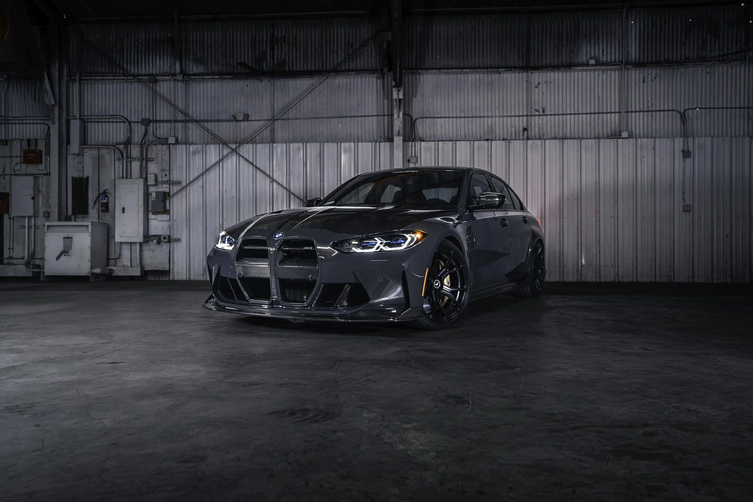 A black BMW M3 sports car parked in an industrial garage with metal walls, dim lighting, and exposed utility boxes.