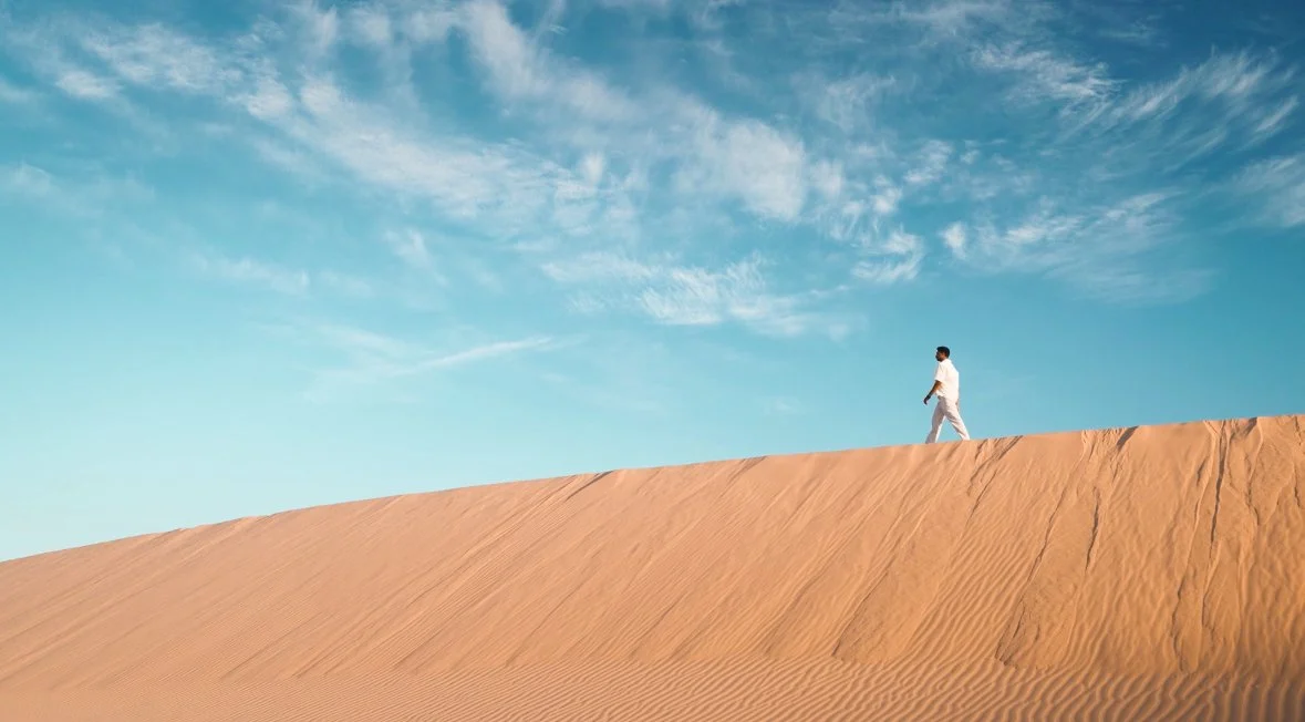 A person wearing white walks on a sand dune in a desert landscape under a blue sky with wispy clouds.